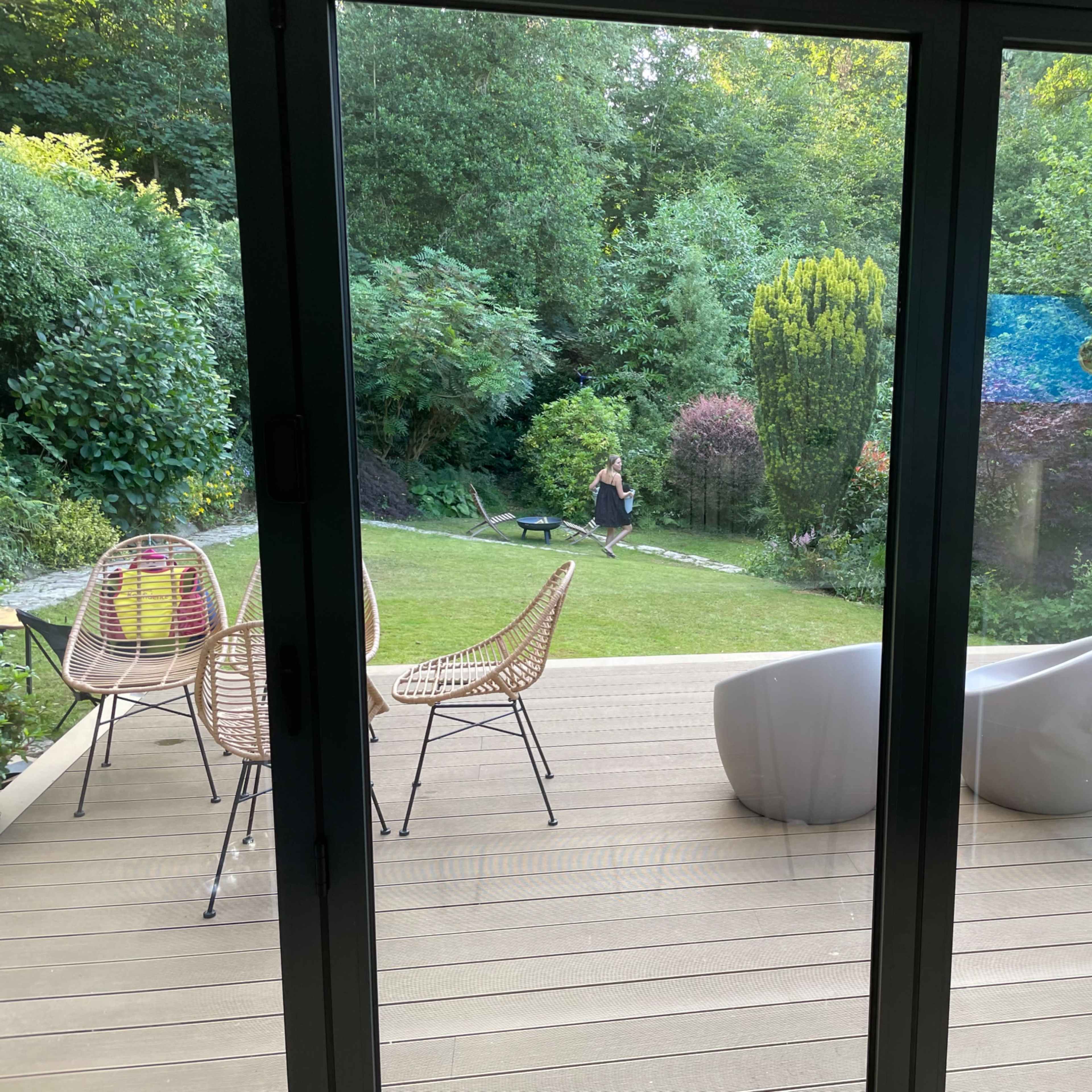 A view from inside a modern home showing a wooden deck, with two woven chairs and a sculptural white seating element facing a grassy garden surrounded by greenery.