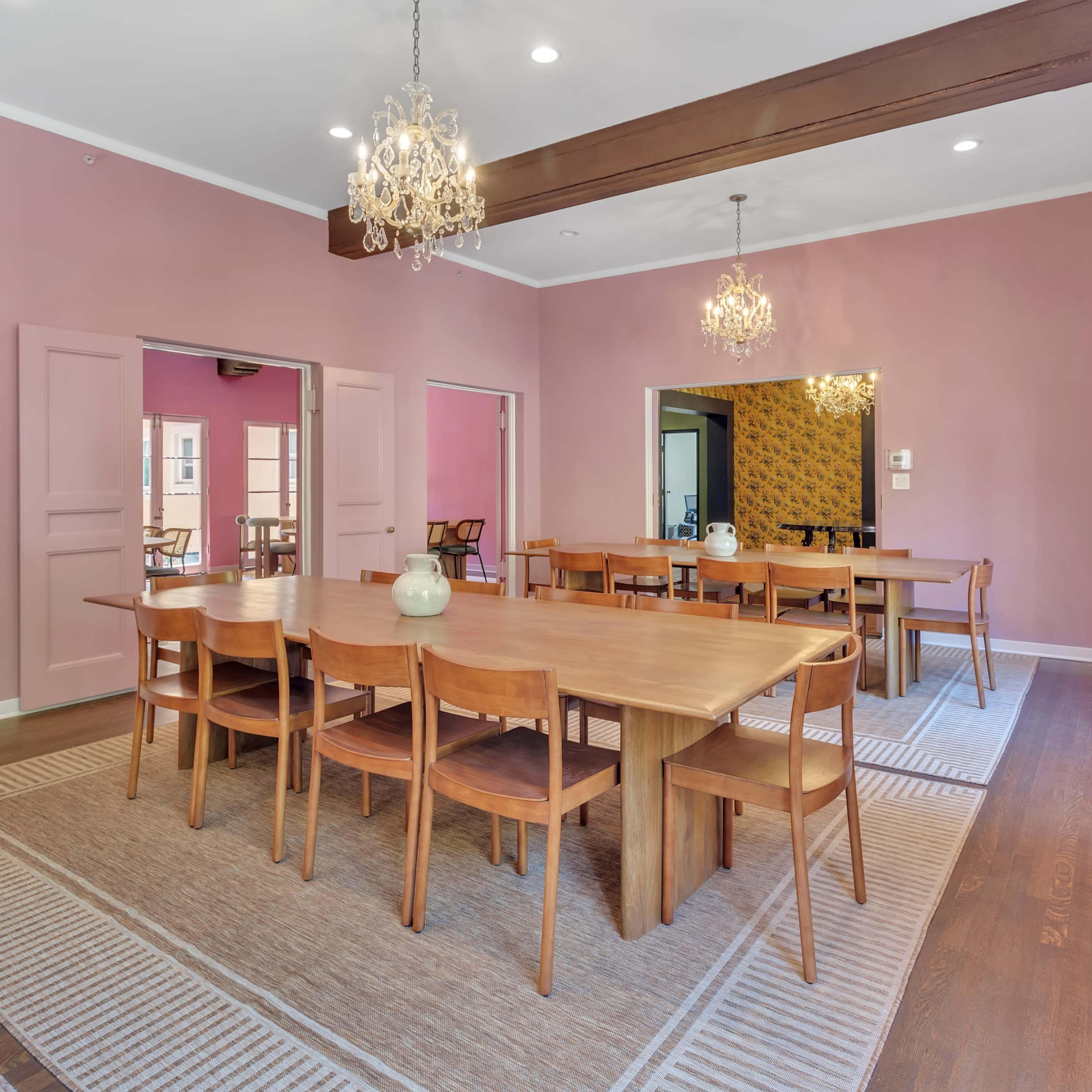 A dining room features a large wooden table surrounded by chairs, with pink walls and chandeliers hanging from the ceiling.