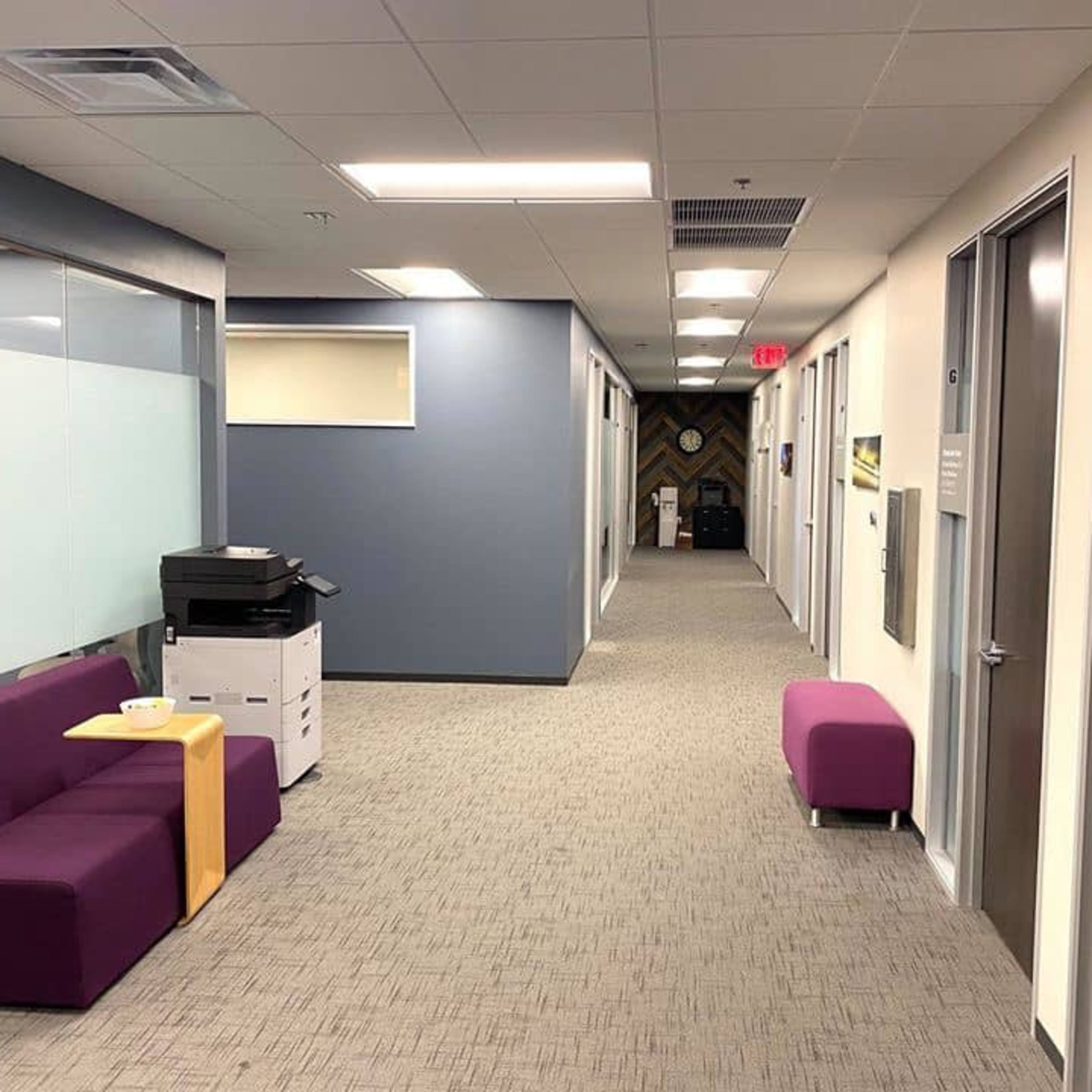 A clean office hallway with purple seating, light gray carpet, and doors leading to individual offices.