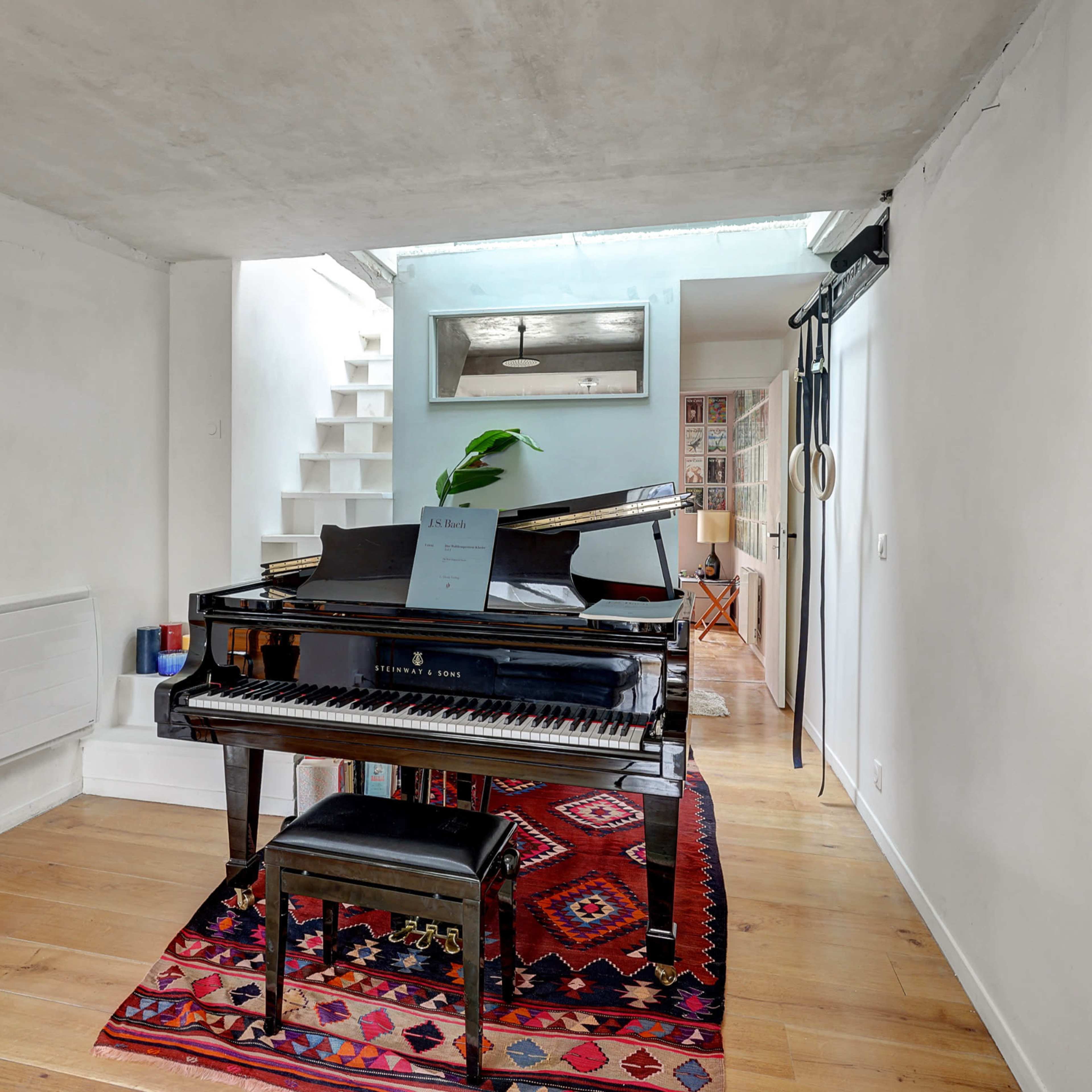 A black piano sits on a colorful rug in a bright room with white walls and a staircase in the background.
