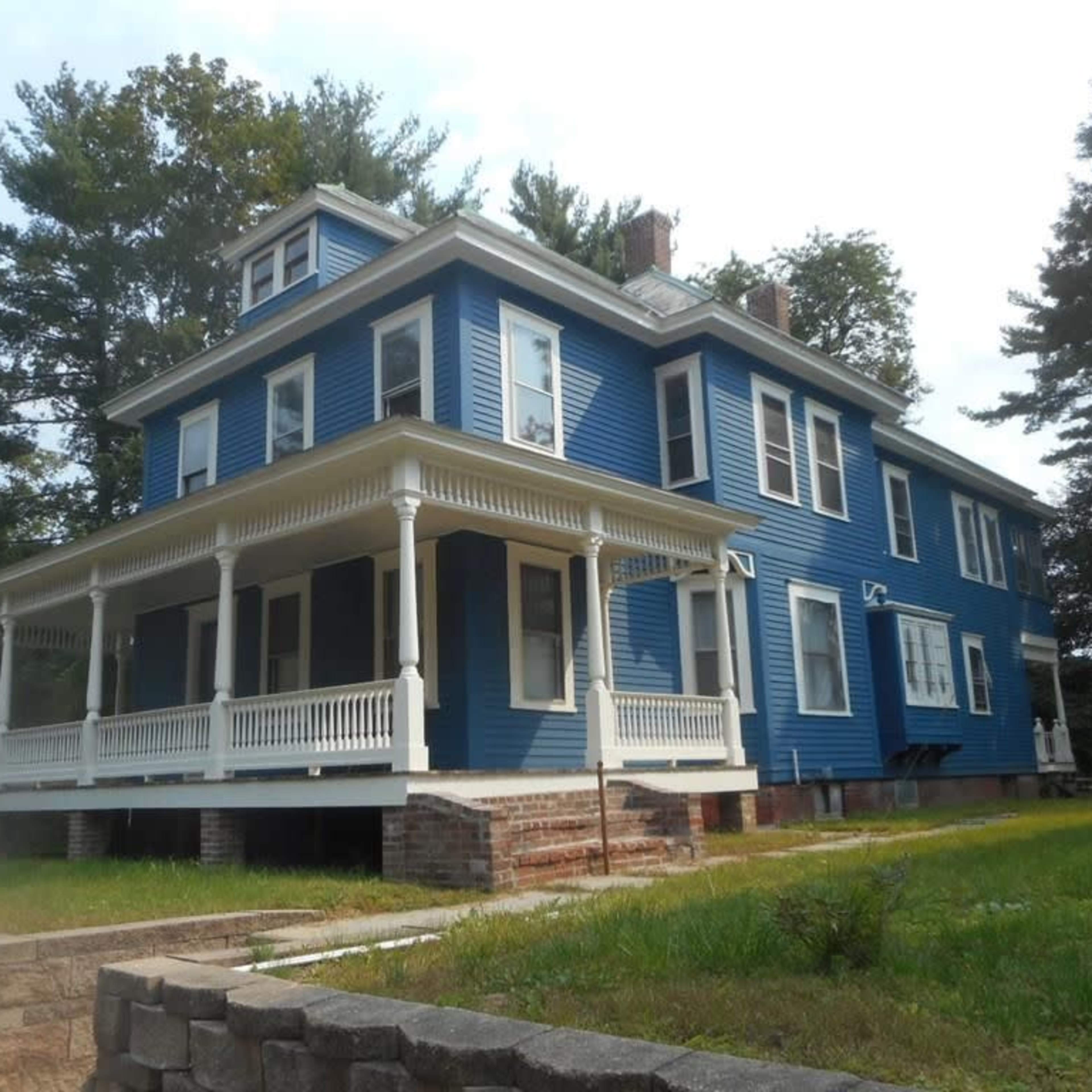 A large blue house with a wraparound porch and multiple gabled roofs, surrounded by trees and a grassy area.