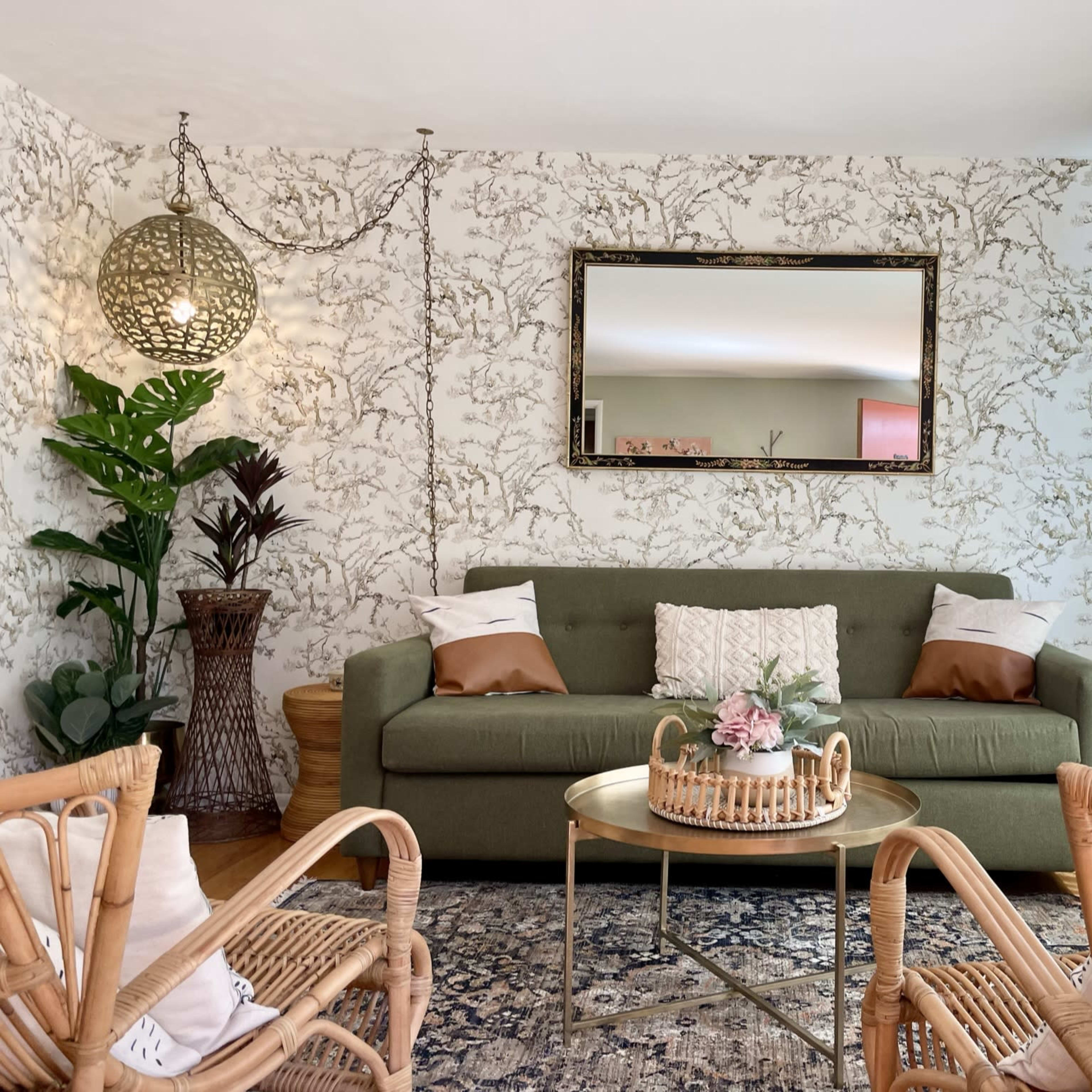 A green sofa with decorative pillows is centered against a patterned wall, surrounded by wicker chairs and a coffee table topped with a small floral arrangement.