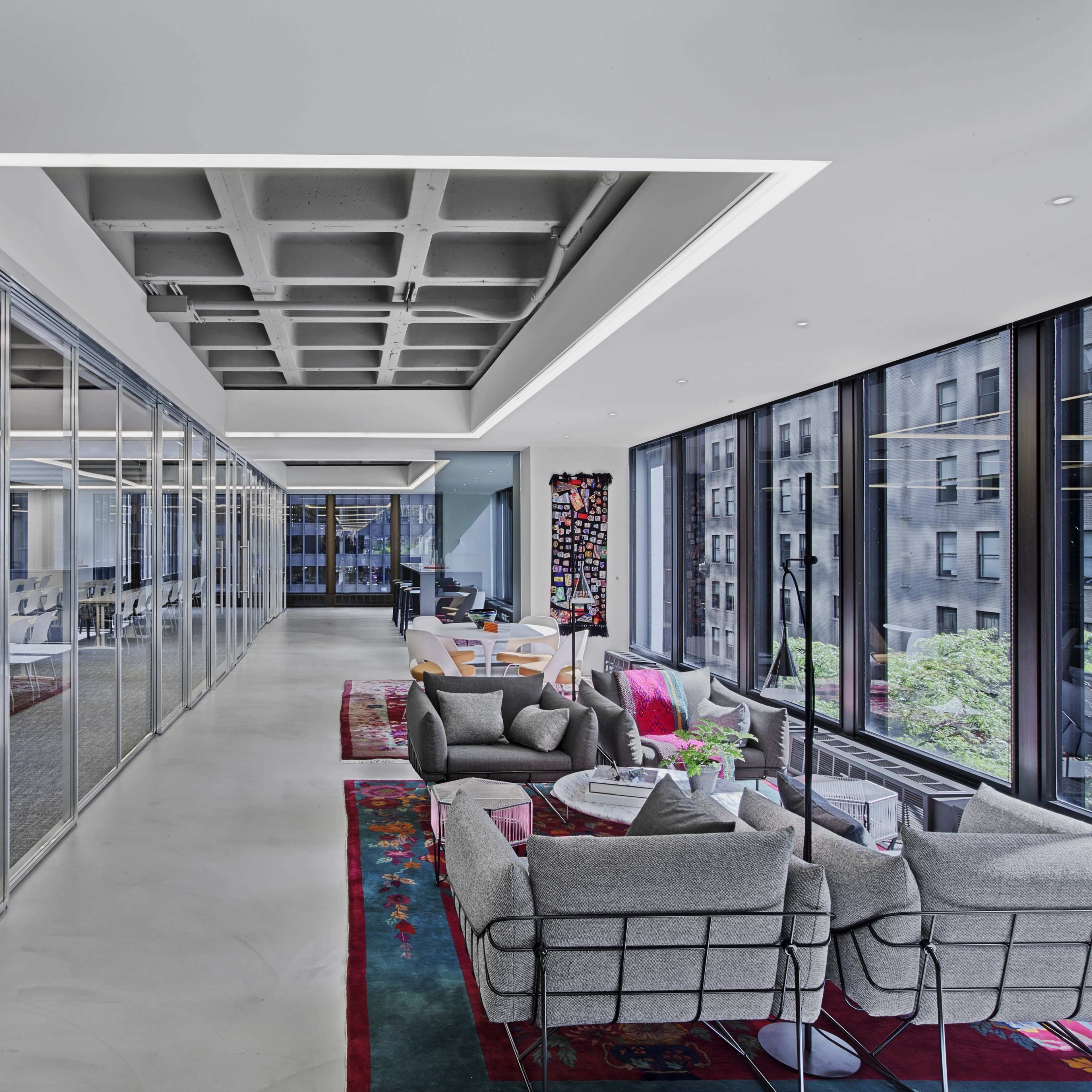 A modern office space with glass walls, a seating area furnished with gray couches, and large windows providing views of the exterior.