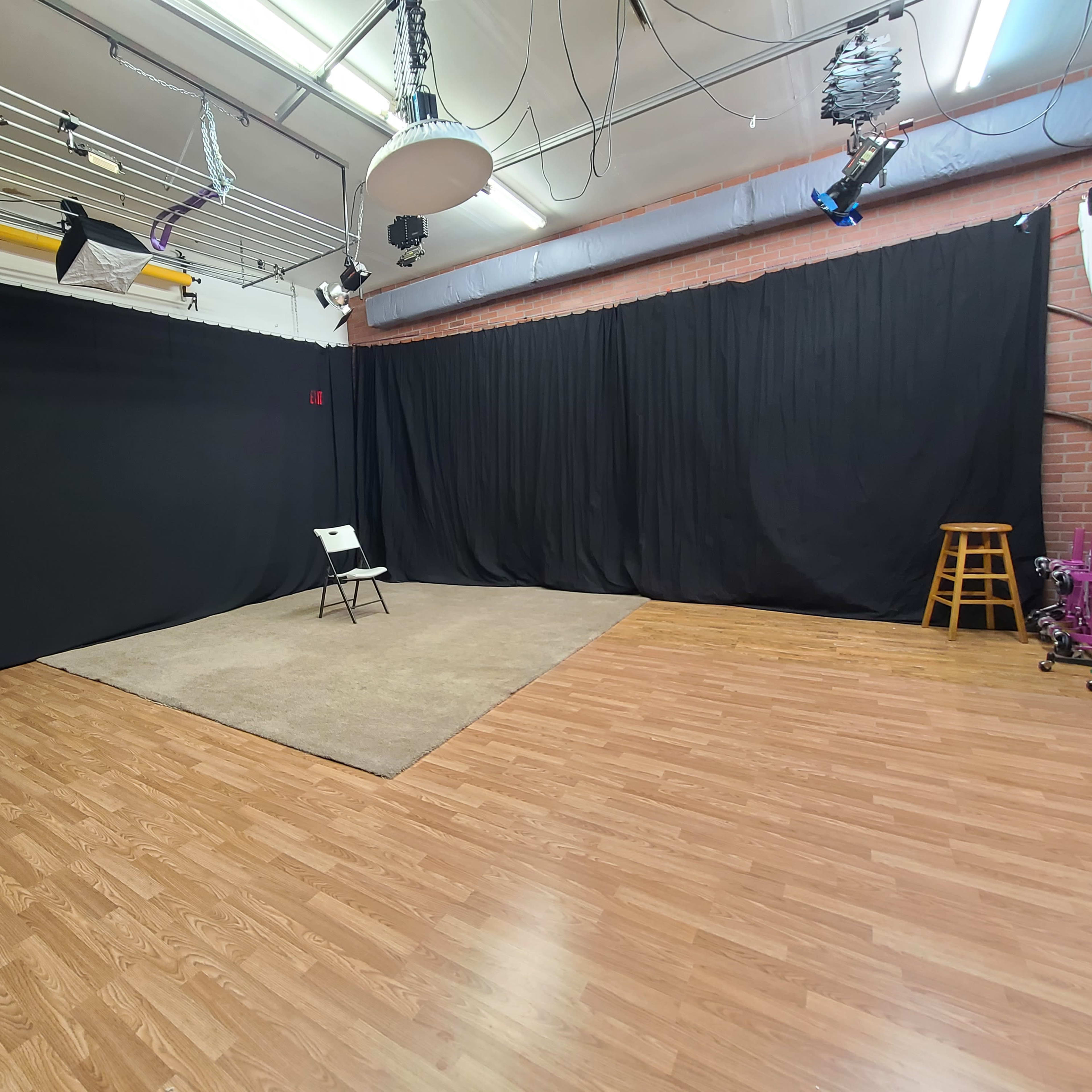 A studio space with black curtains, a beige rug, a single chair in the center, and wooden flooring.