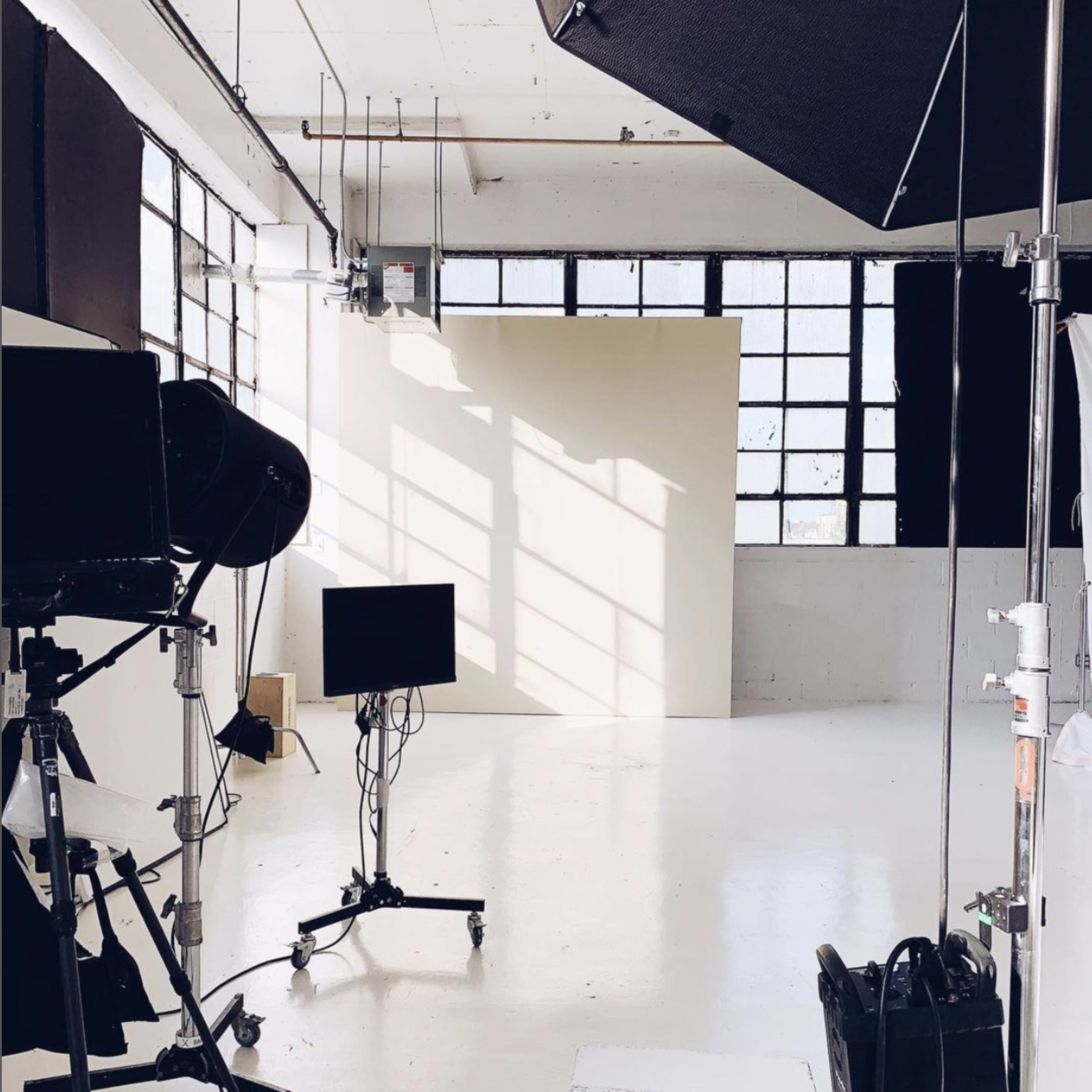 A photography studio with a blank backdrop, lighting equipment, and a monitor set up under large windows.
