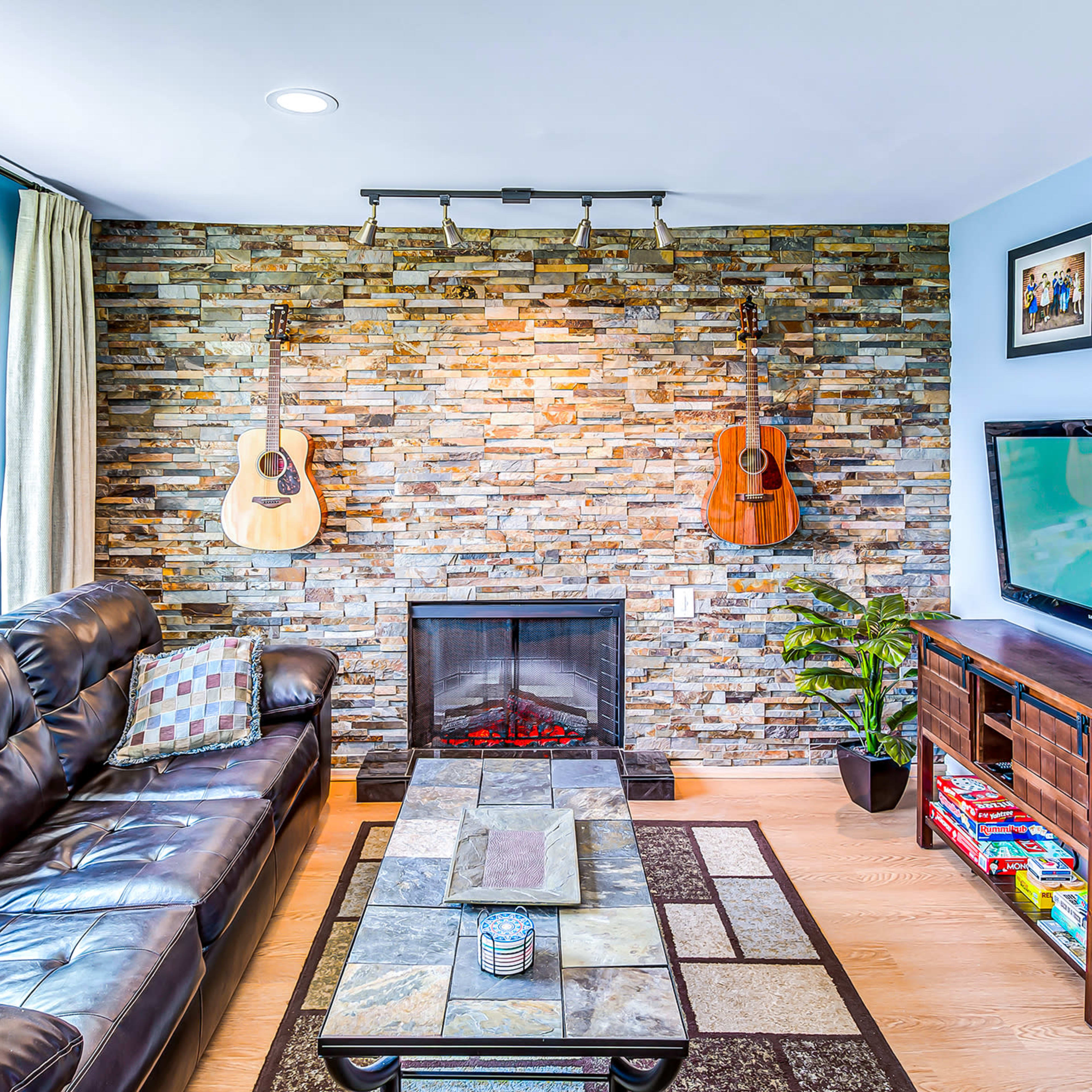 A living room with a stone feature wall, two acoustic guitars hanging on the wall, a black leather couch, a modern television, and a decorative coffee table.