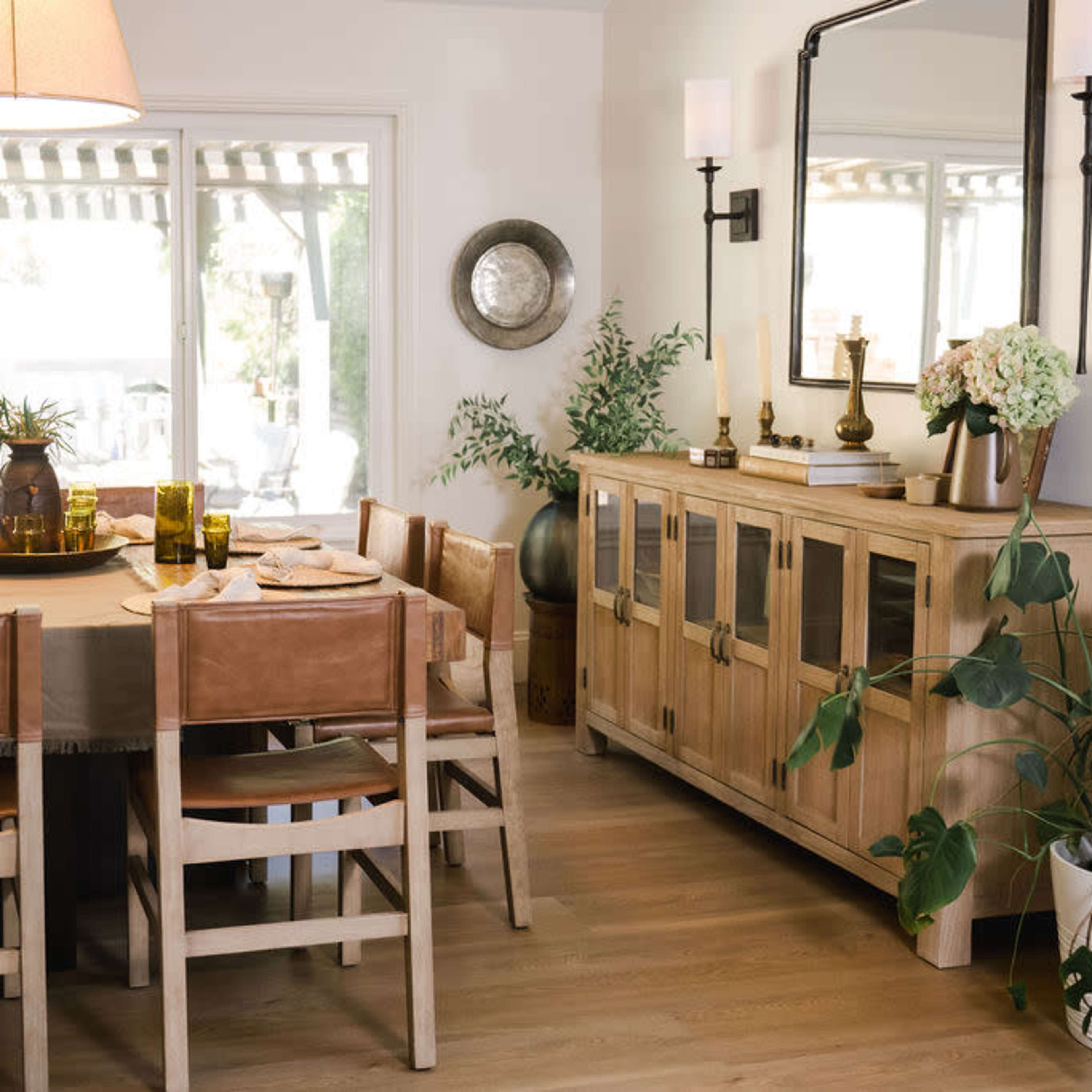 A dining room features a large wooden table surrounded by leather chairs, a sideboard with glass doors, and plants enhancing the decor.