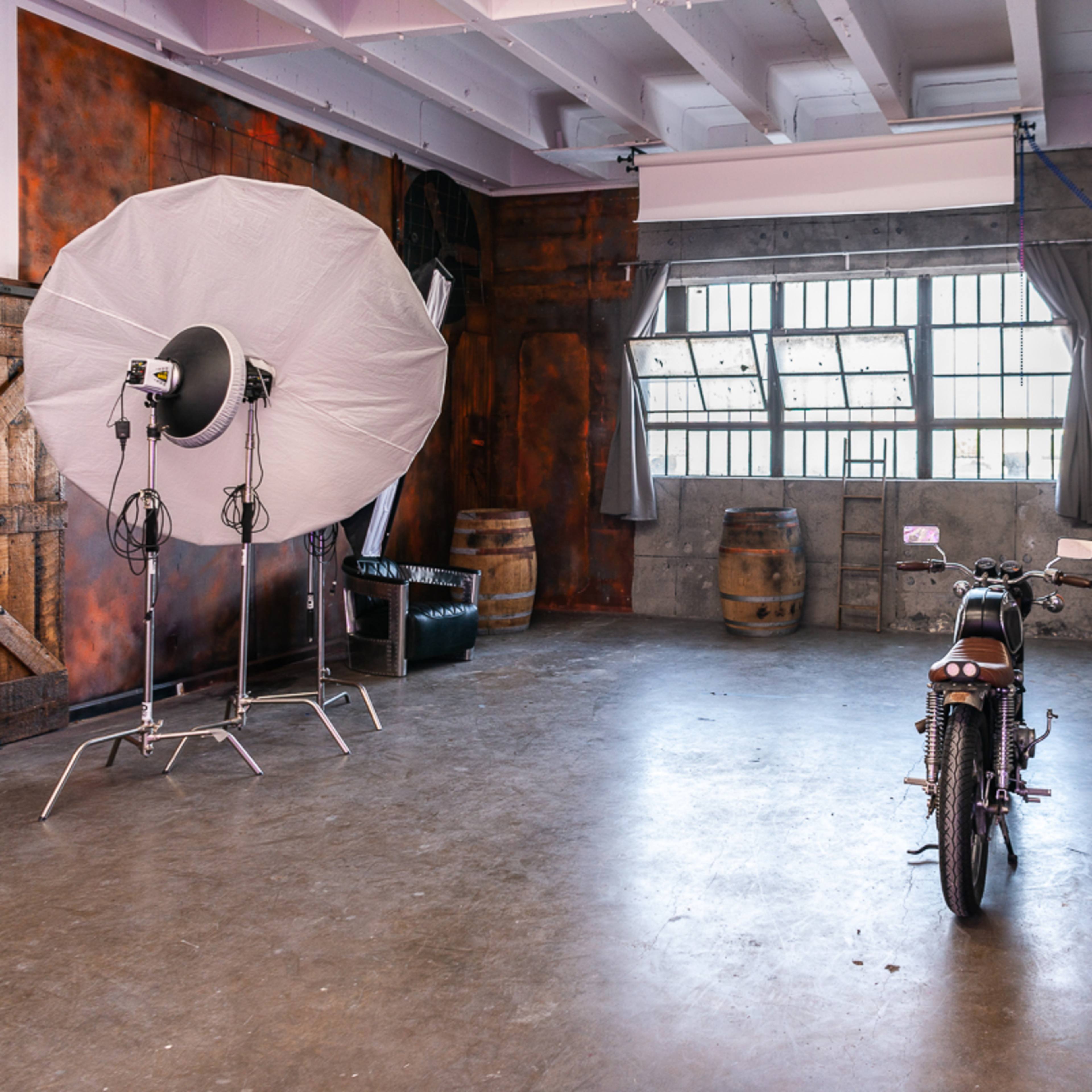 A spacious studio interior featuring a large softbox light, a vintage motorcycle, wooden barrels, and industrial-style walls.