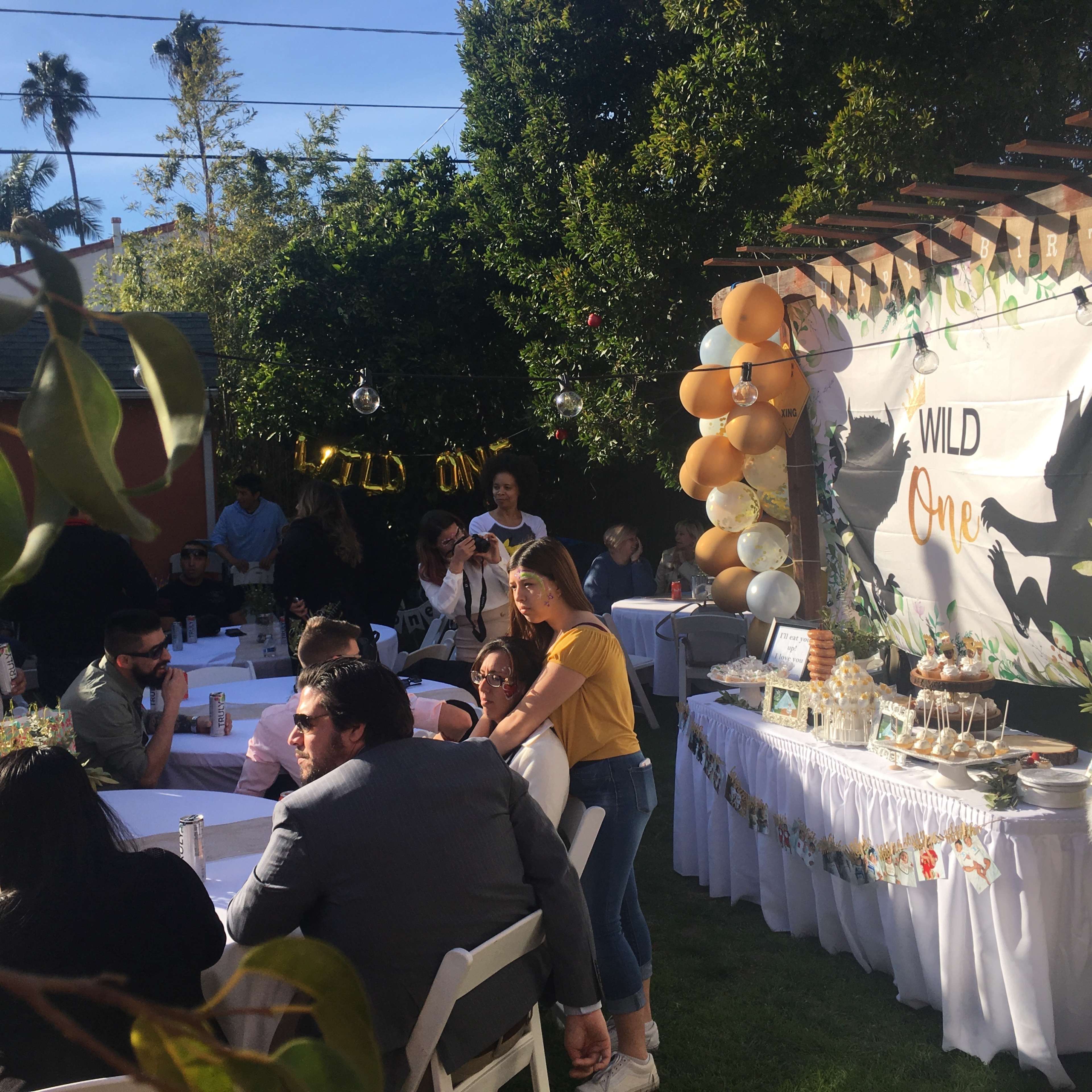 A lively outdoor party scene features guests sitting at white tables amidst greenery, with a decorated backdrop that reads "Wild One."