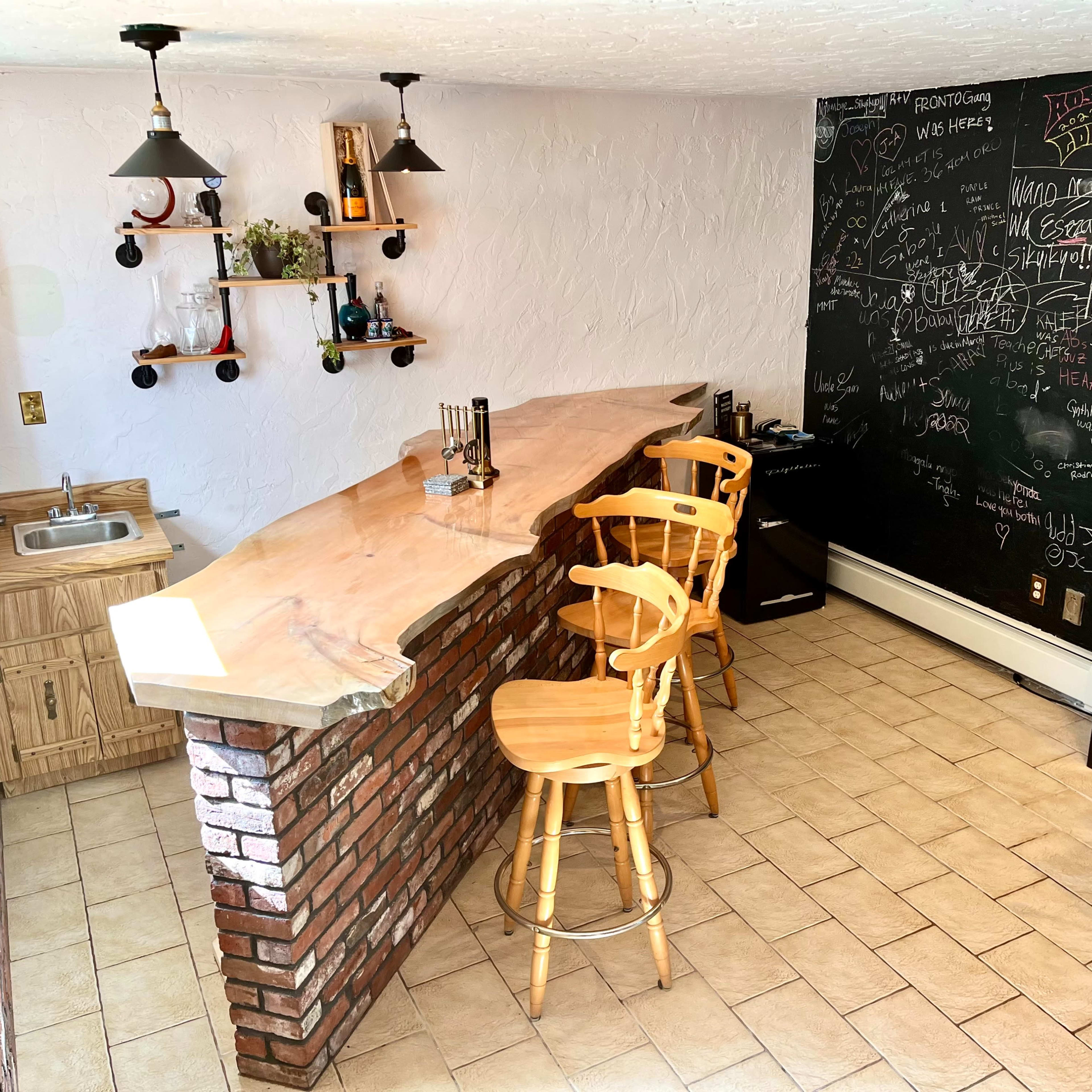 A home bar area featuring a wooden counter with three high stools, a sink to the left, a black chalkboard wall covered in writing, and shelves displaying various bottles.