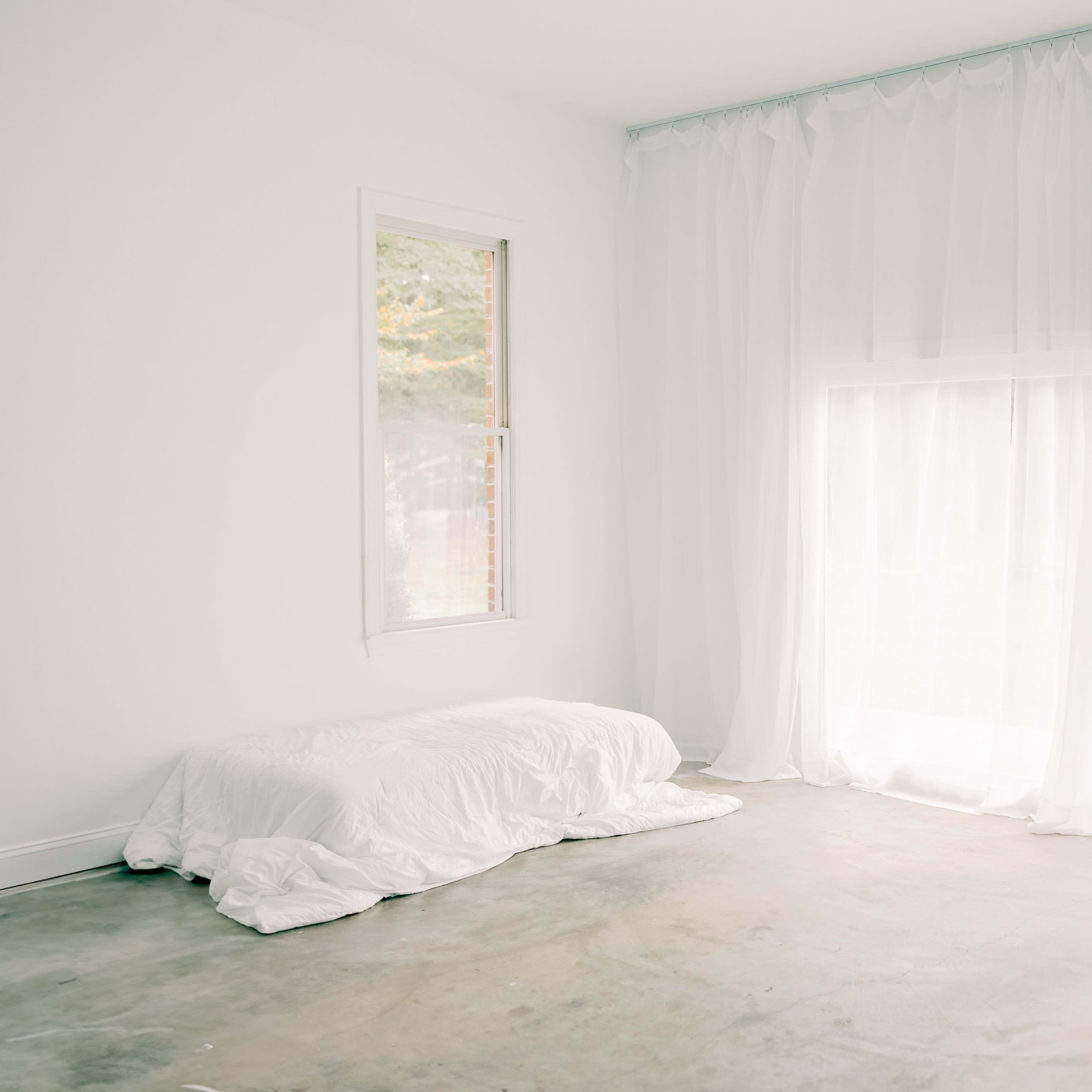 A minimalistic room with a bed covered in white linens and sheer curtains allowing light to filter in through a large window.