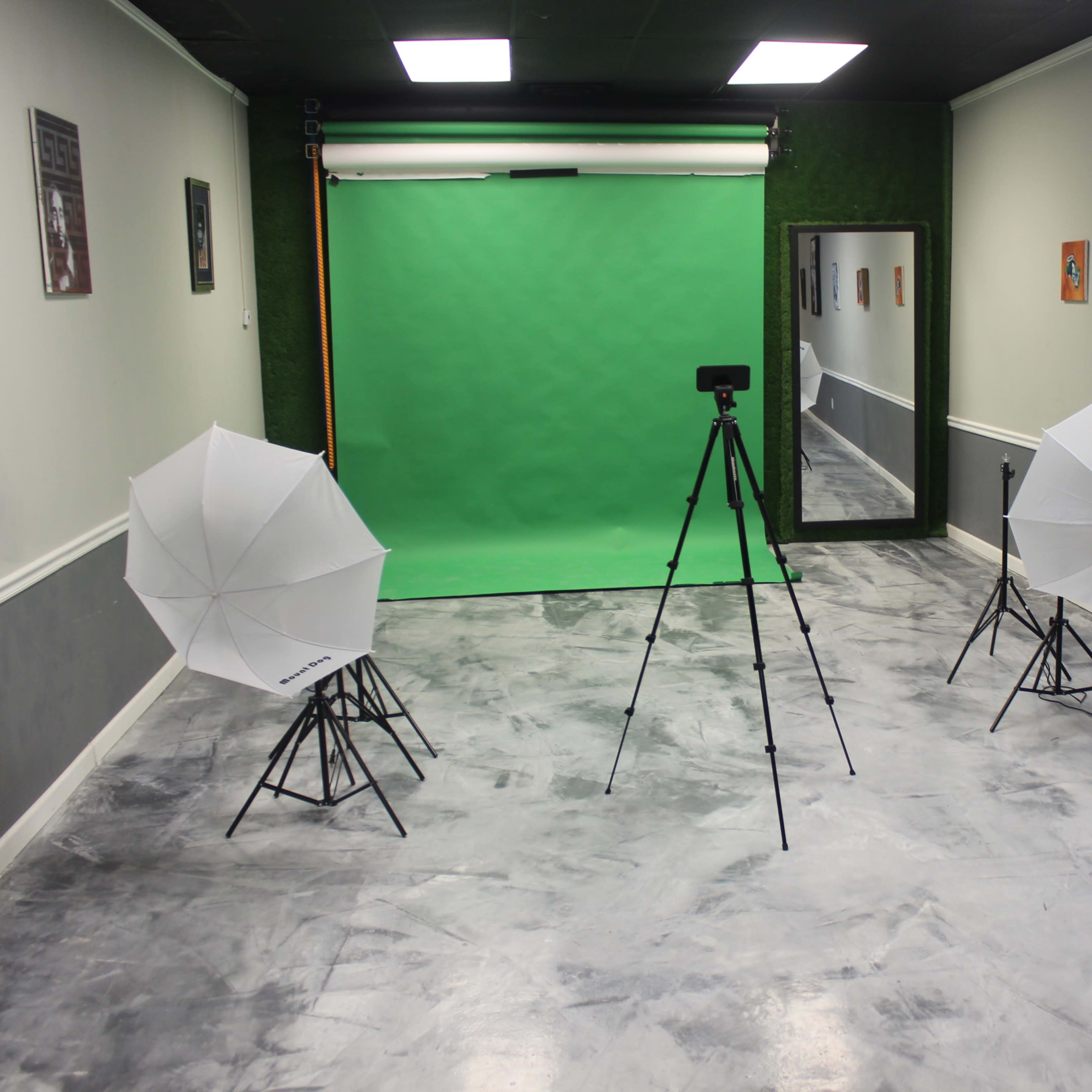 A photography studio setup with a green backdrop, two softbox lights on tripods, and a camera on a tripod in a sparsely decorated room.