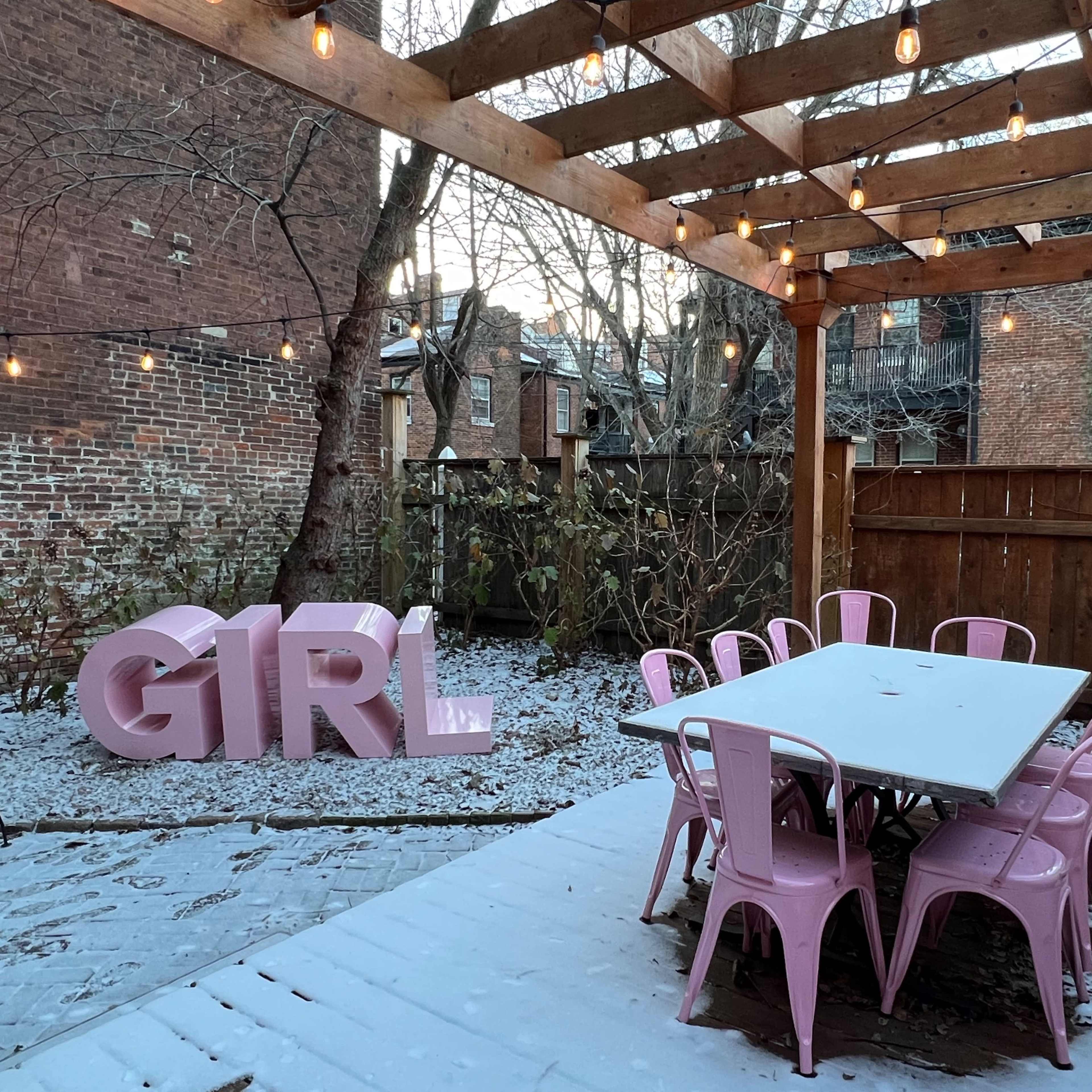 A pink "GIRL" sign rests on the snowy ground near a wooden table and chairs under a pergola with string lights.