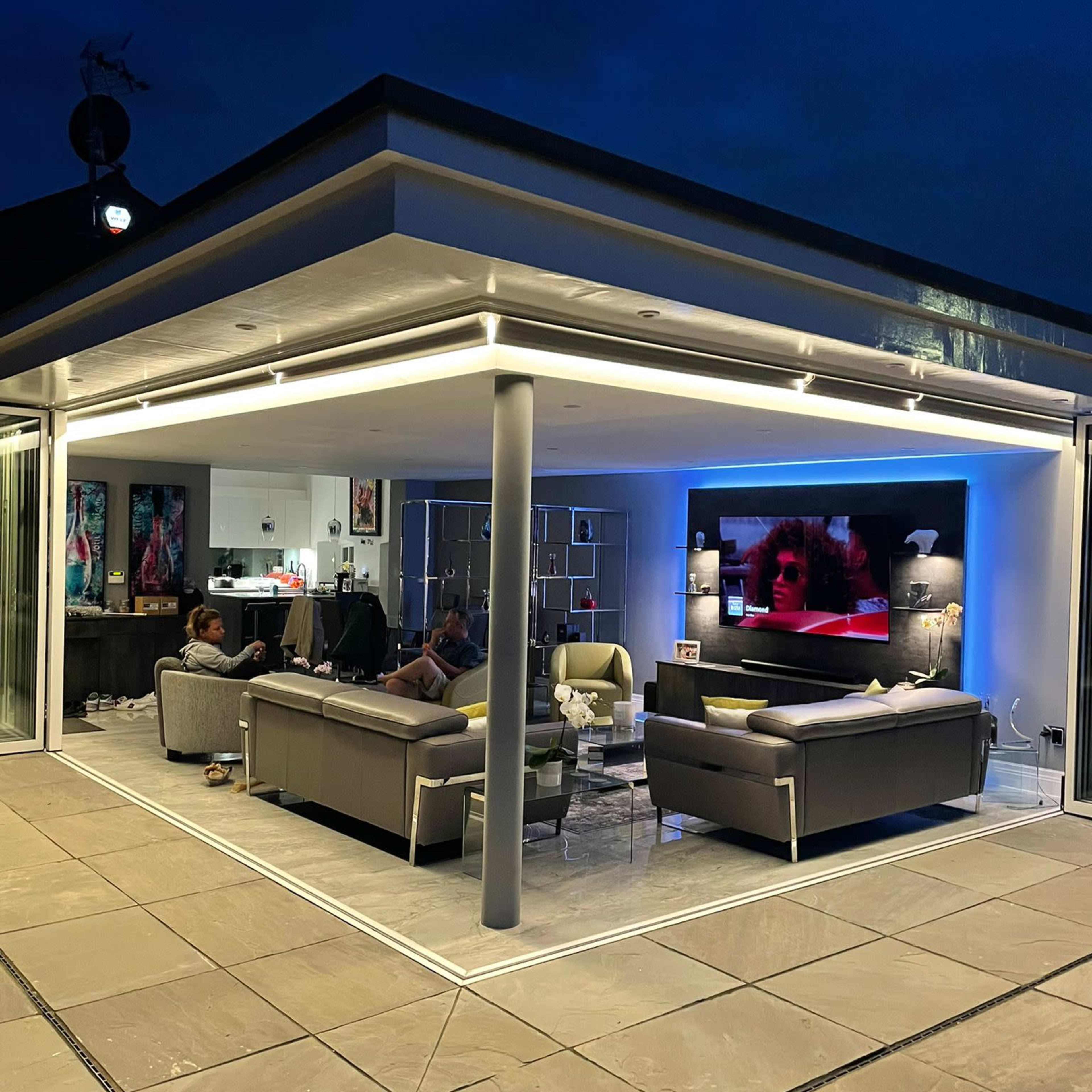 A modern, open-plan living area at night with a large television displaying a scene on one wall and comfortable seating arranged around it.