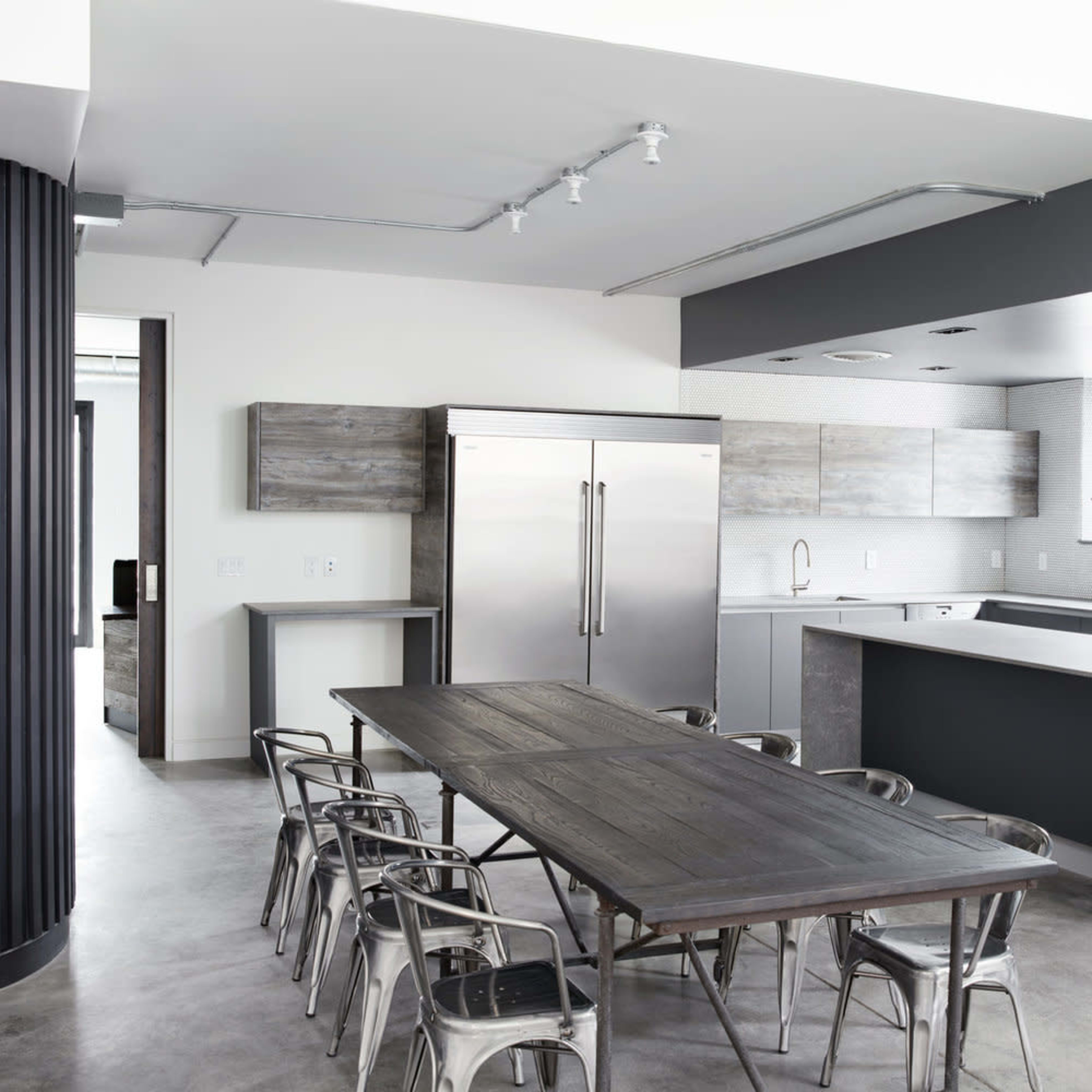 A modern kitchen features stainless steel appliances, gray cabinetry, and a long wooden dining table surrounded by metal chairs.