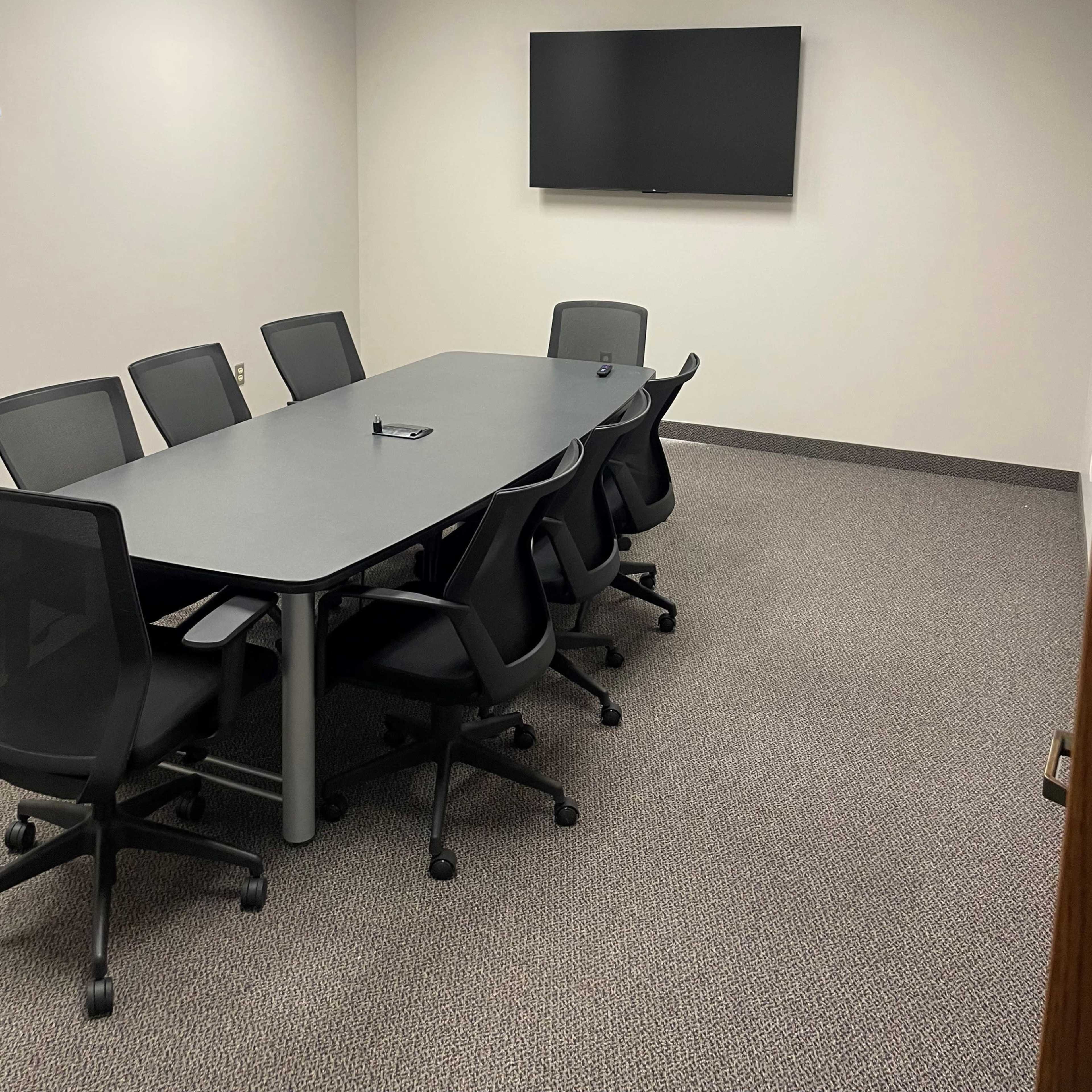 A conference room features a rectangular table surrounded by six swivel chairs and a wall-mounted flat-screen TV.