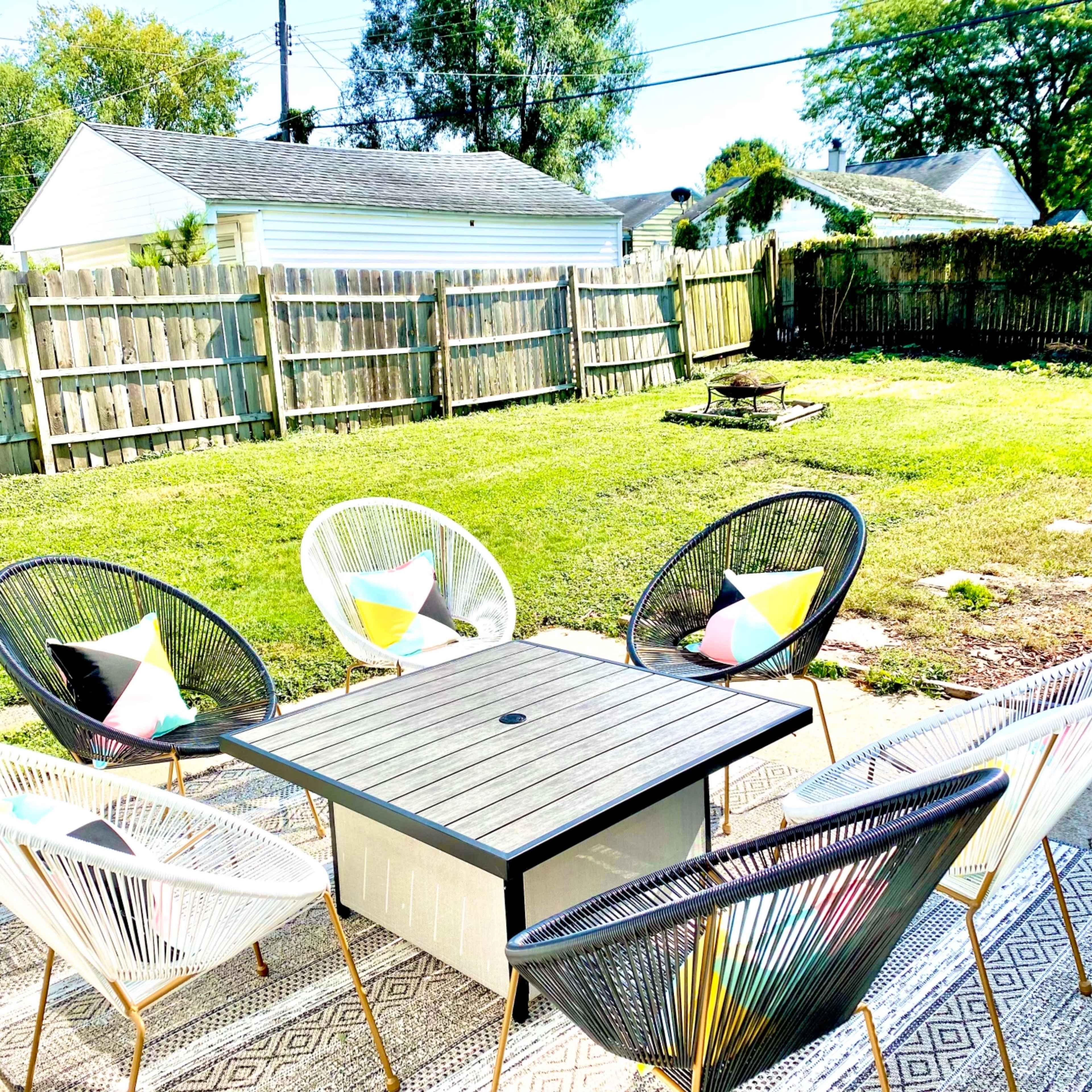 A patio area features a square table surrounded by four chairs and a rug, set against a grassy yard enclosed by wooden fencing.
