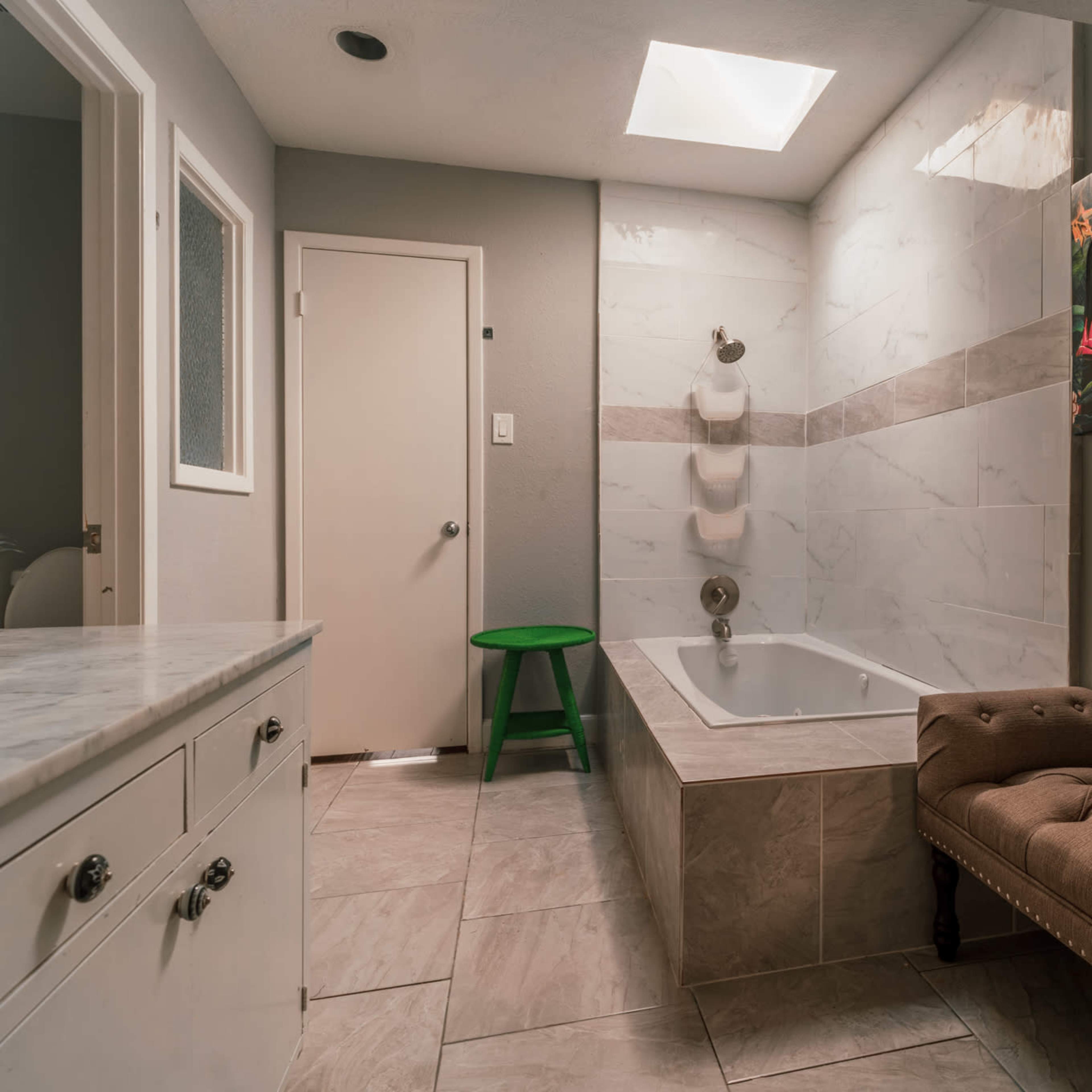 A bathroom with a bathtub, a patterned artwork on the wall, a green stool, and a vanity with drawers.