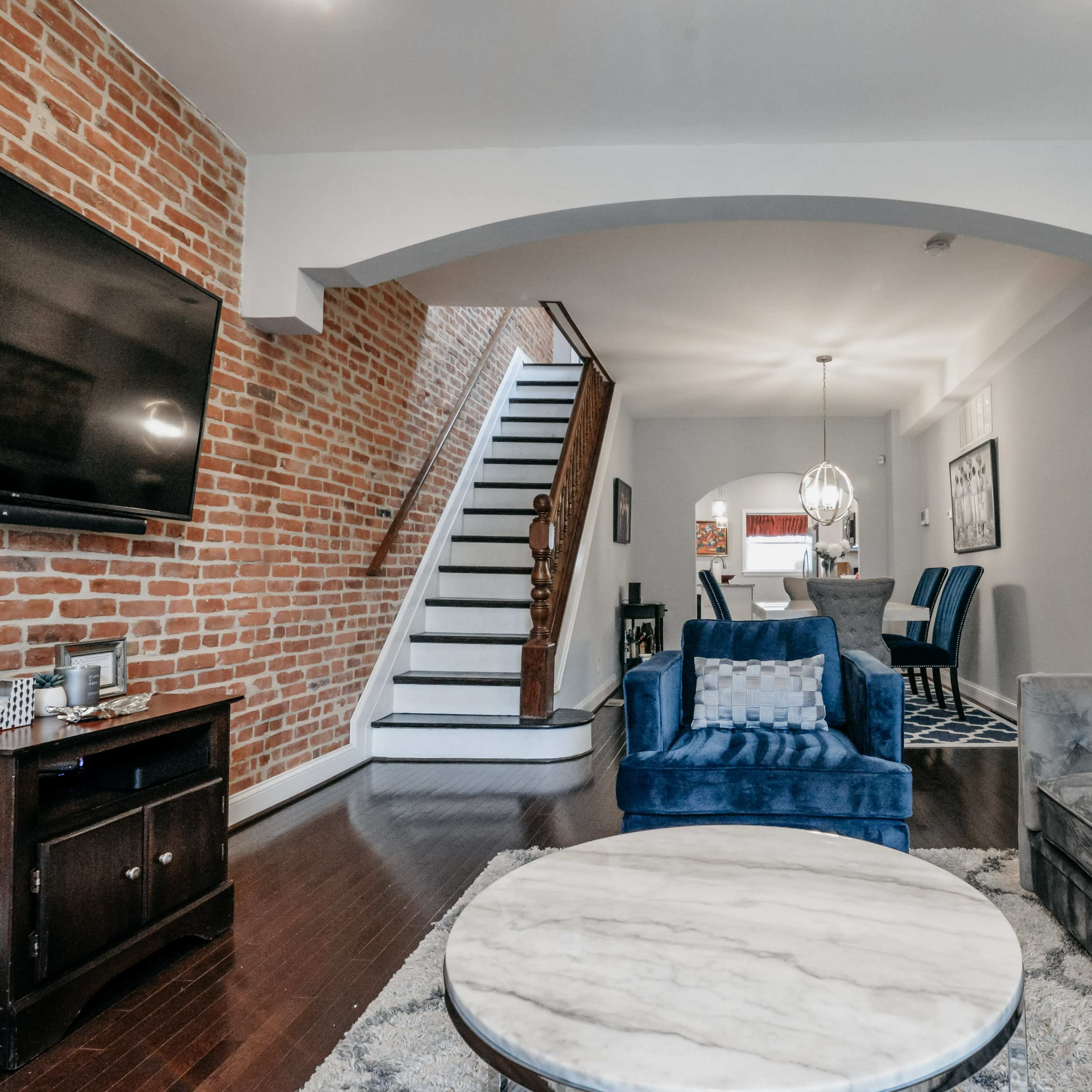 A living room featuring a blue sofa, a marble coffee table, a large television, and an exposed brick wall, with a staircase leading to an upper level in the background.