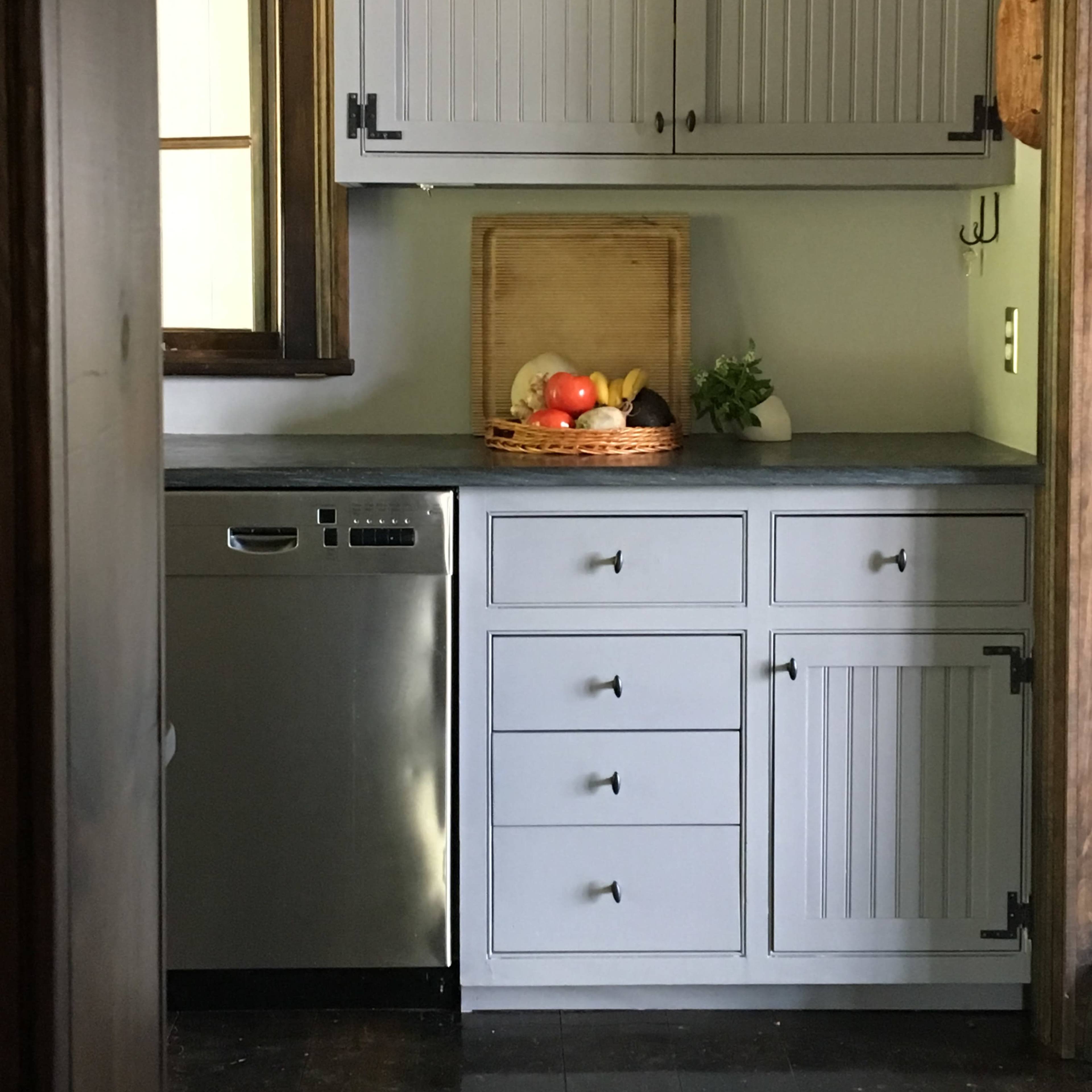 A kitchen features gray cabinets, a stainless steel dishwasher, and a fruit basket on the countertop.