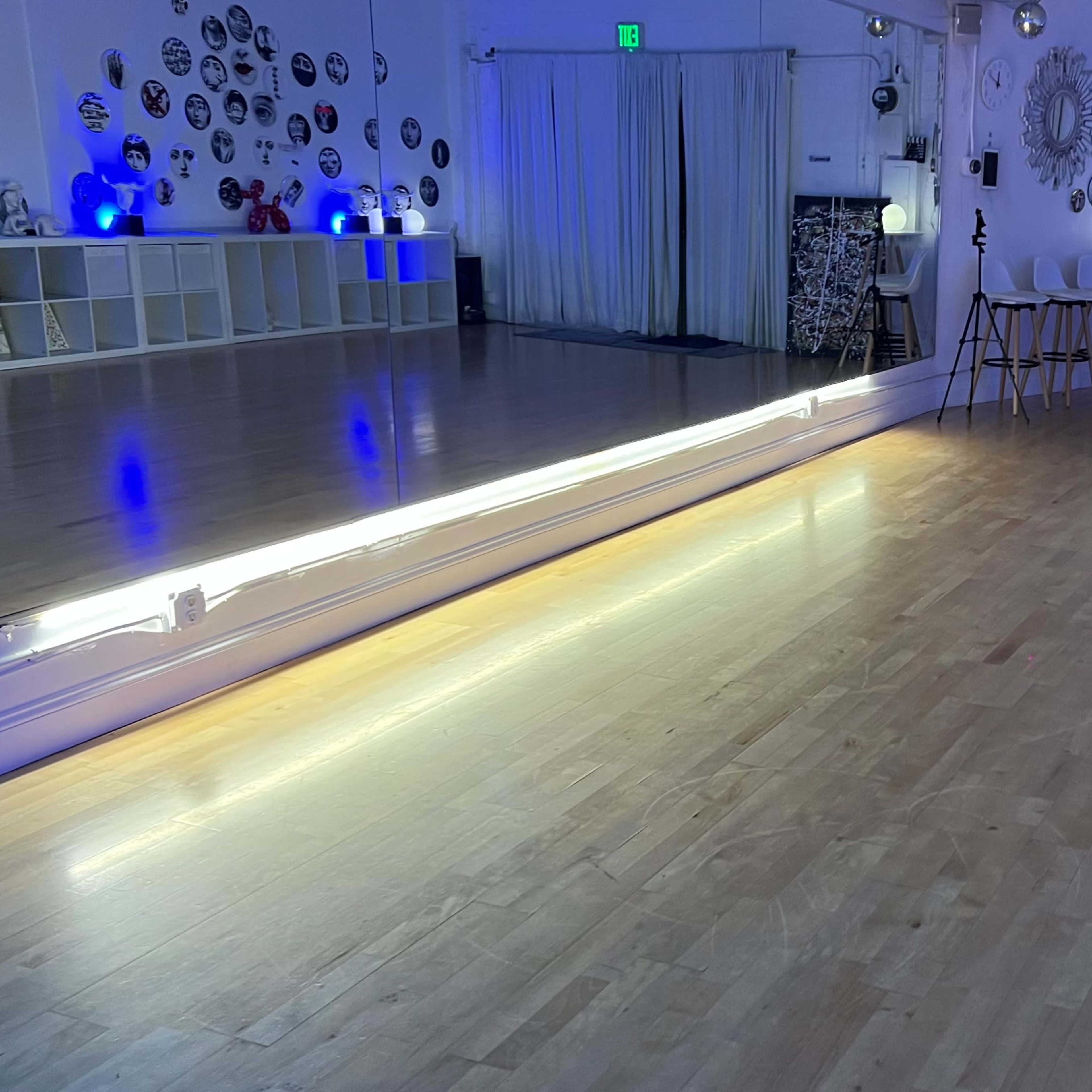 A dance studio with a wooden floor, a large mirror, and blue lighting, along with a seating area featuring three chairs.