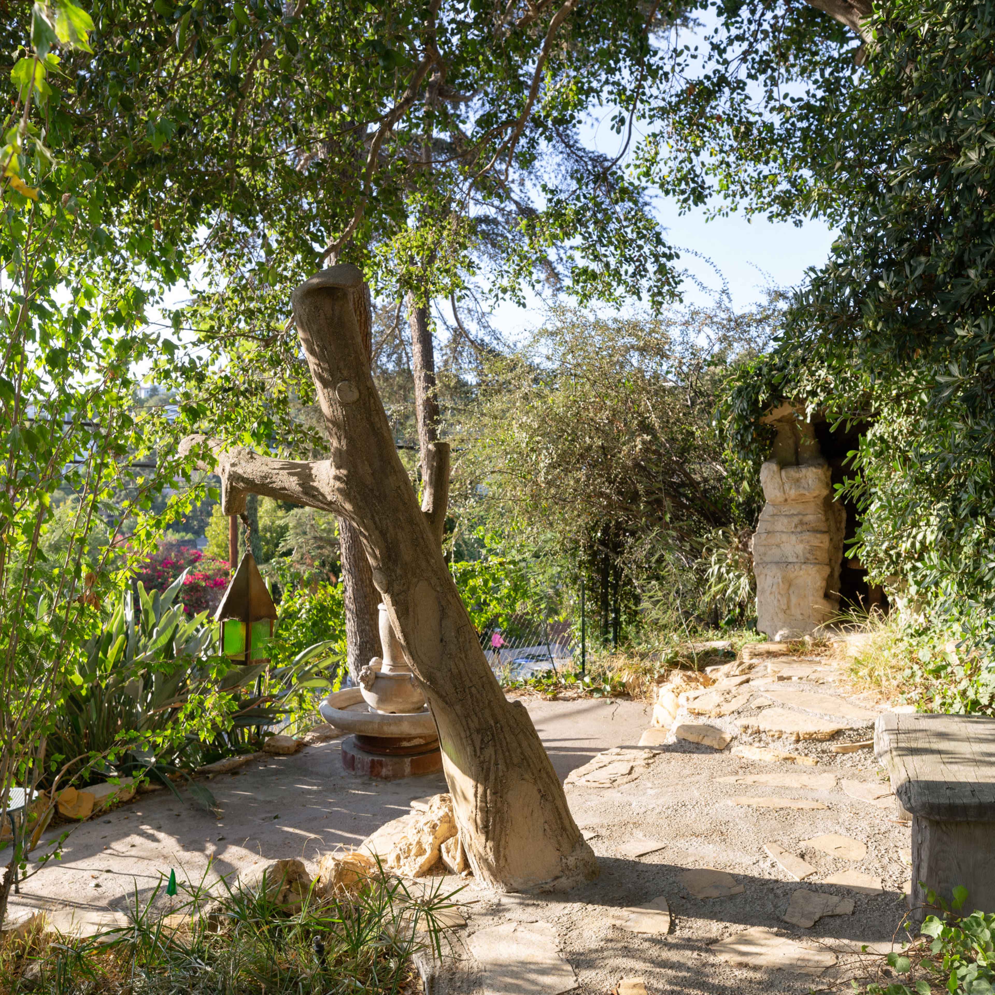 A tranquil garden area featuring a leaning tree, stone pathways, and lush greenery surrounding a small seating area.