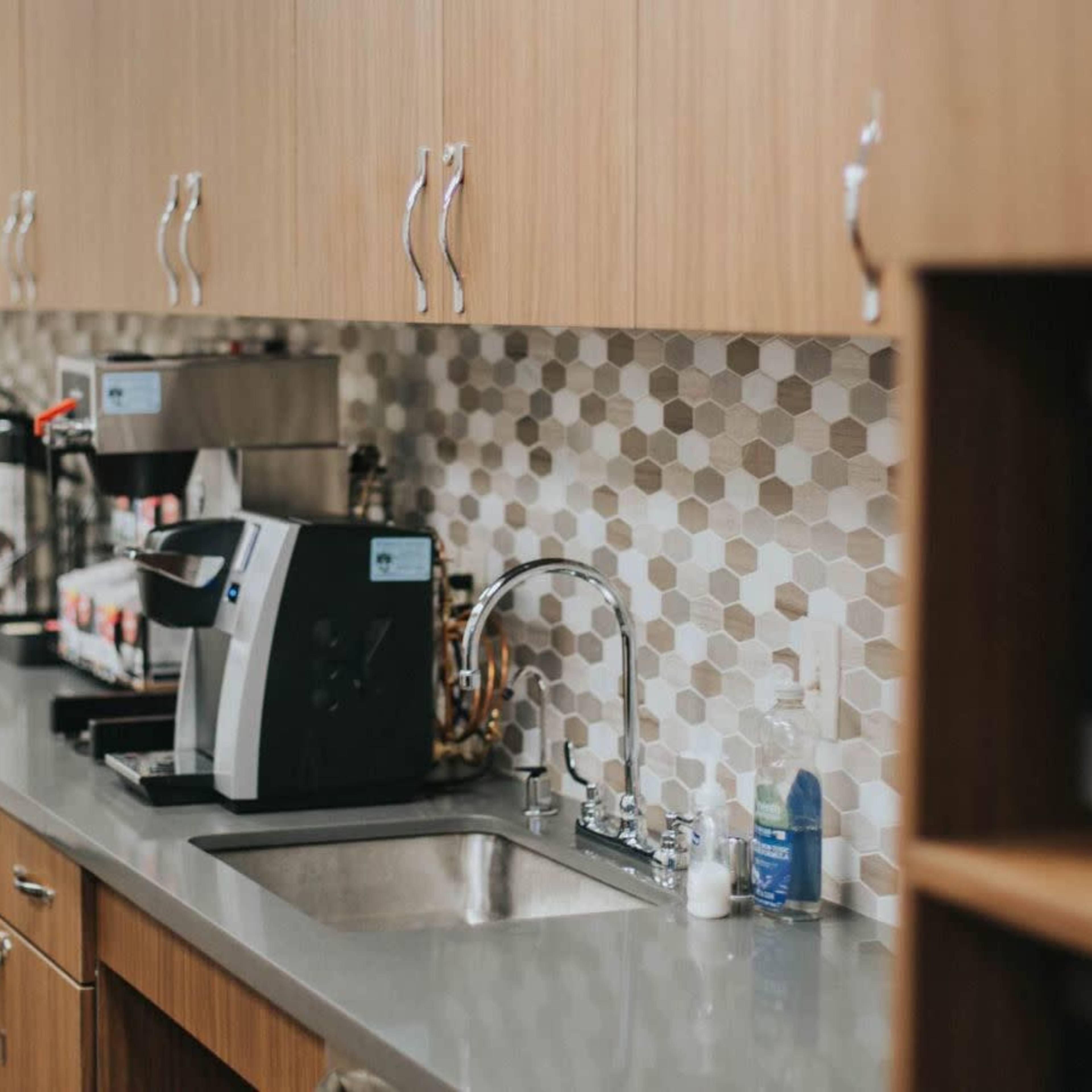 A modern kitchen area with a countertop, a sink, coffee machines, and a patterned backsplash.