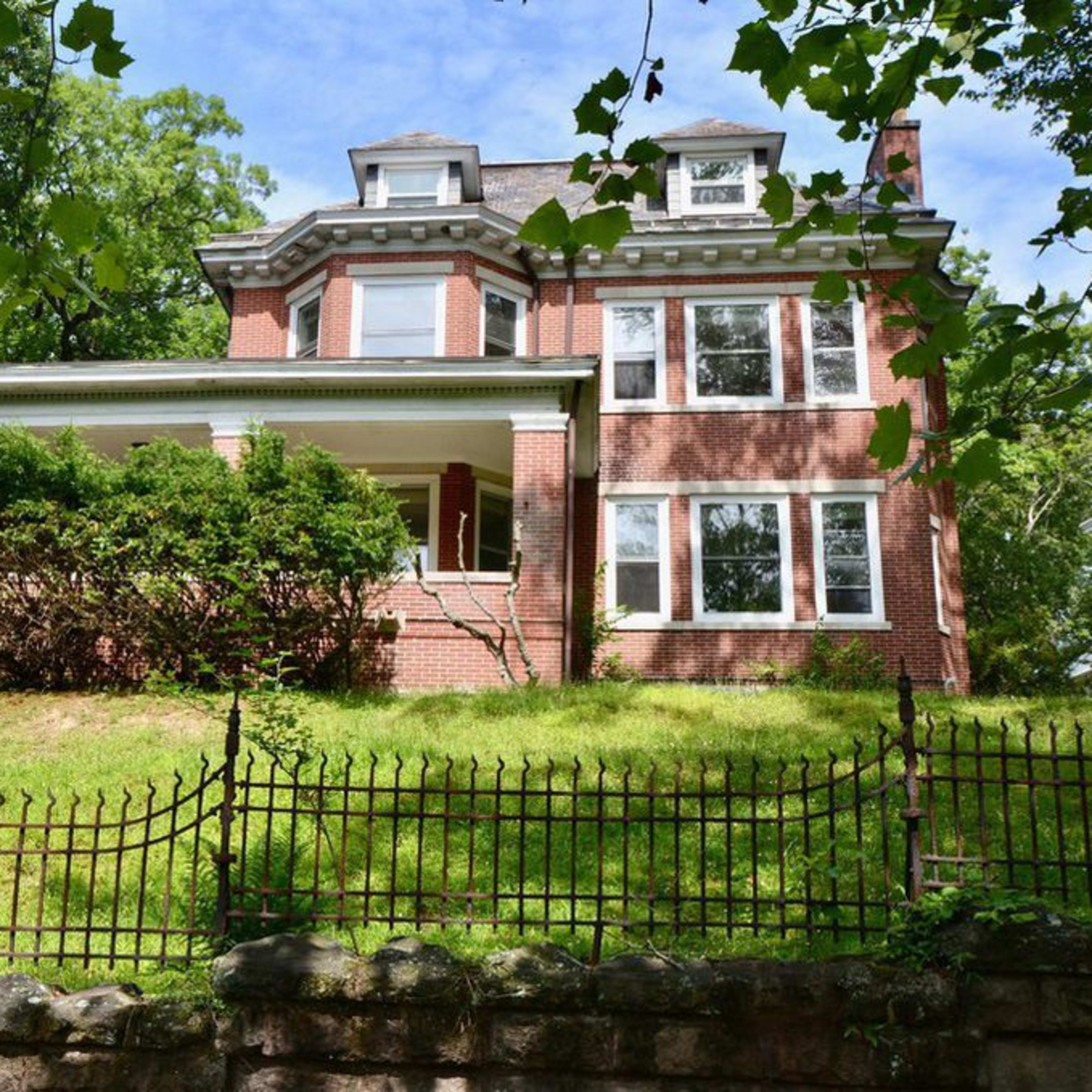 A brick house with multiple windows and a porch is situated on a grassy lot enclosed by an iron fence.