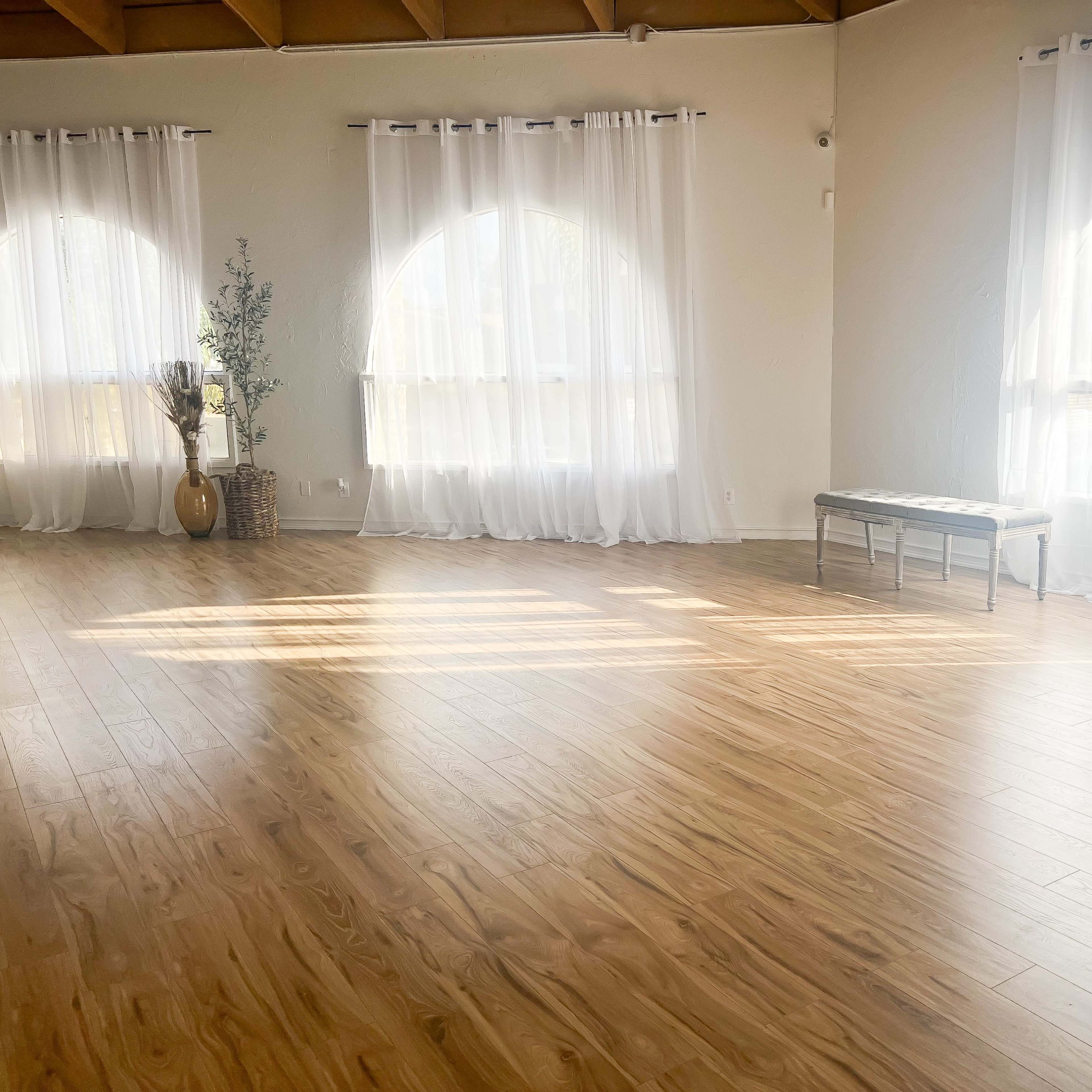 A bright room with large arched windows, sheer white curtains, a wooden floor, and a decorative vase beside a bench.