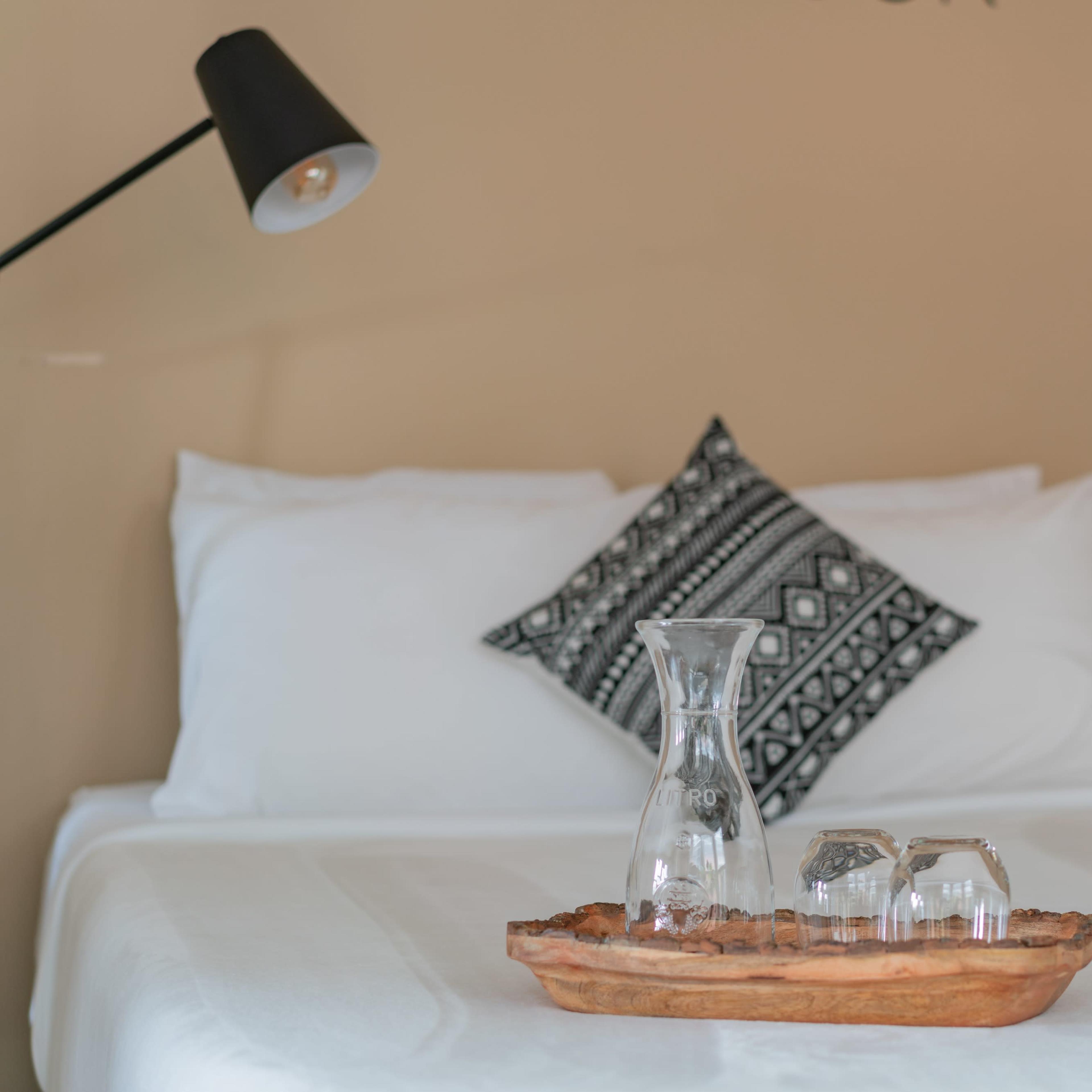 A glass decanter and two glasses are placed on a wooden tray on a neatly made bed with a patterned pillow.