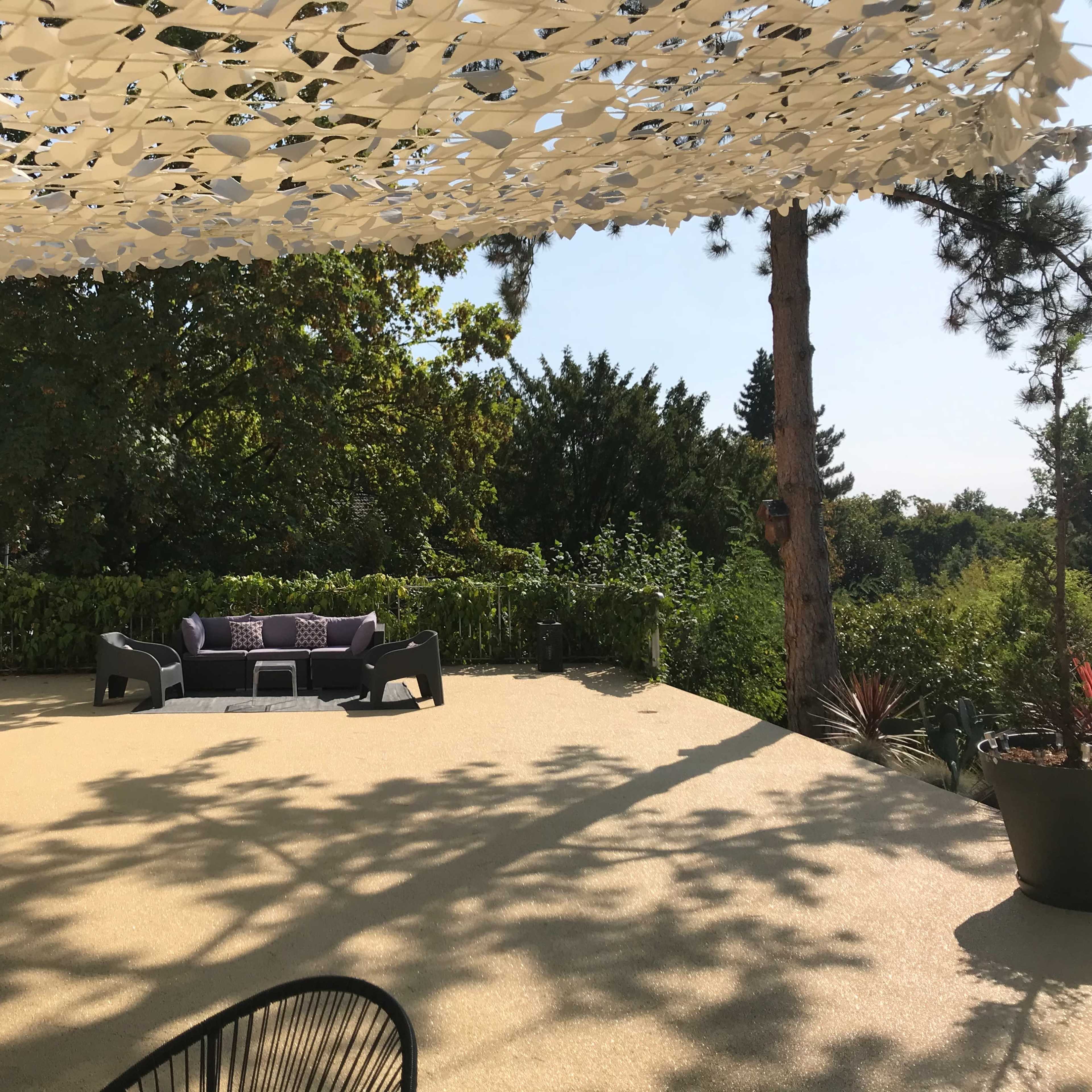 A shaded outdoor patio features a seating area with dark furniture surrounded by greenery.