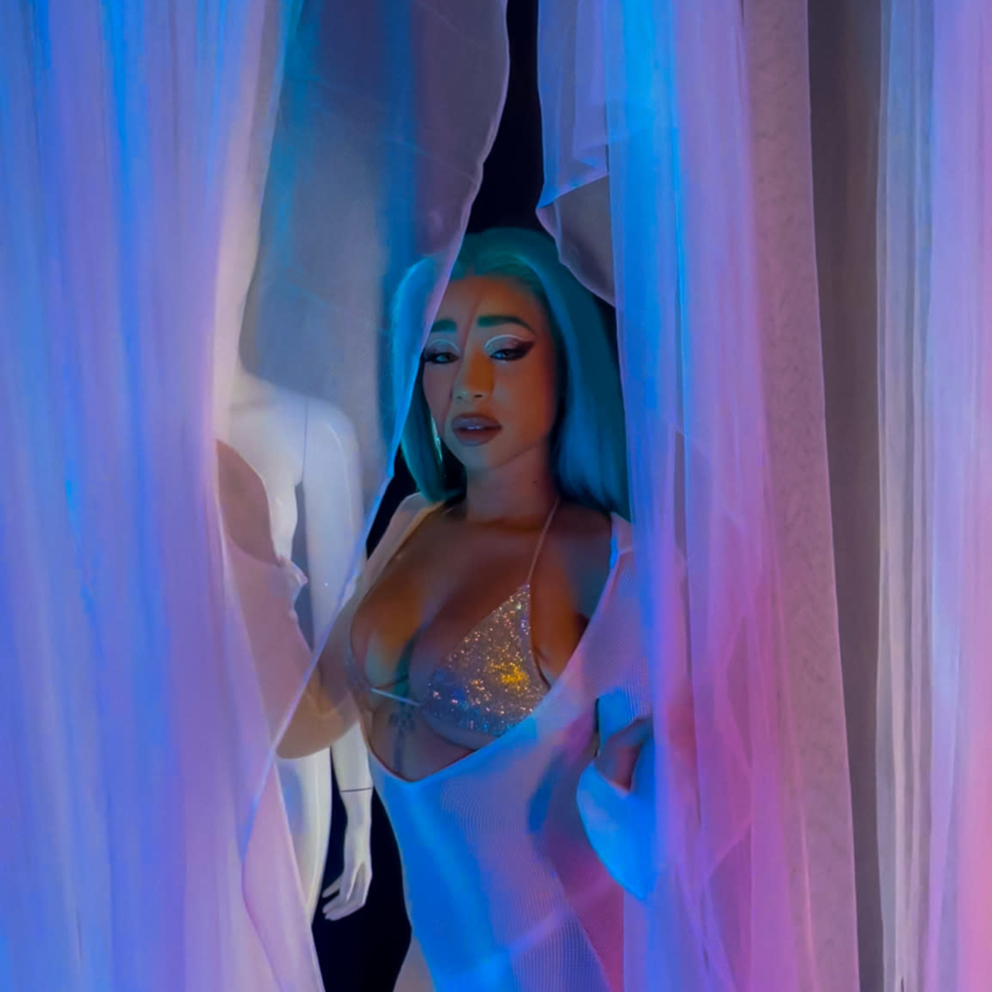 A woman with turquoise hair stands between sheer curtains illuminated with blue and pink light.