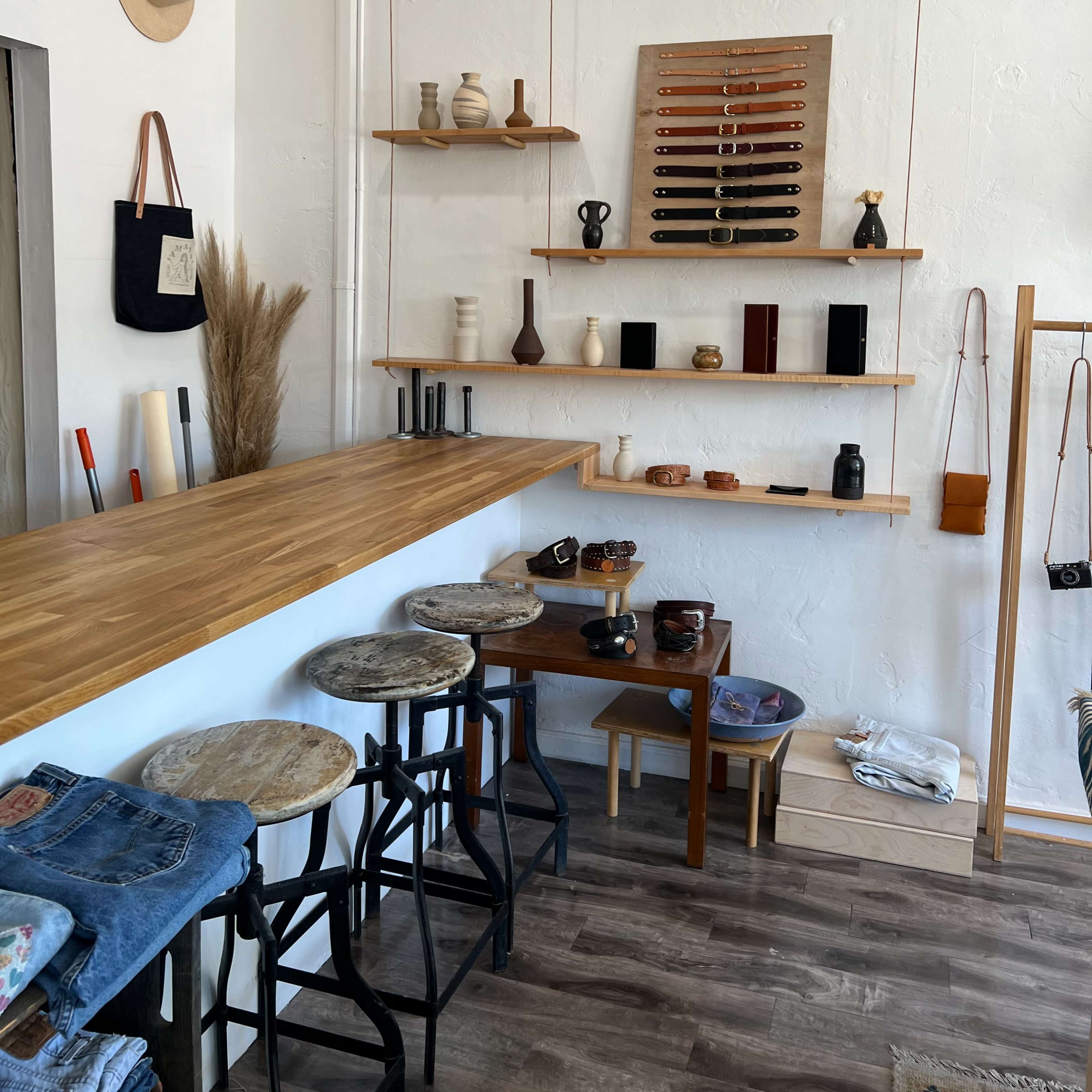 An interior of a retail space featuring wooden shelves displaying various items, a bar counter with stools, and a section for clothing arranged on the floor.