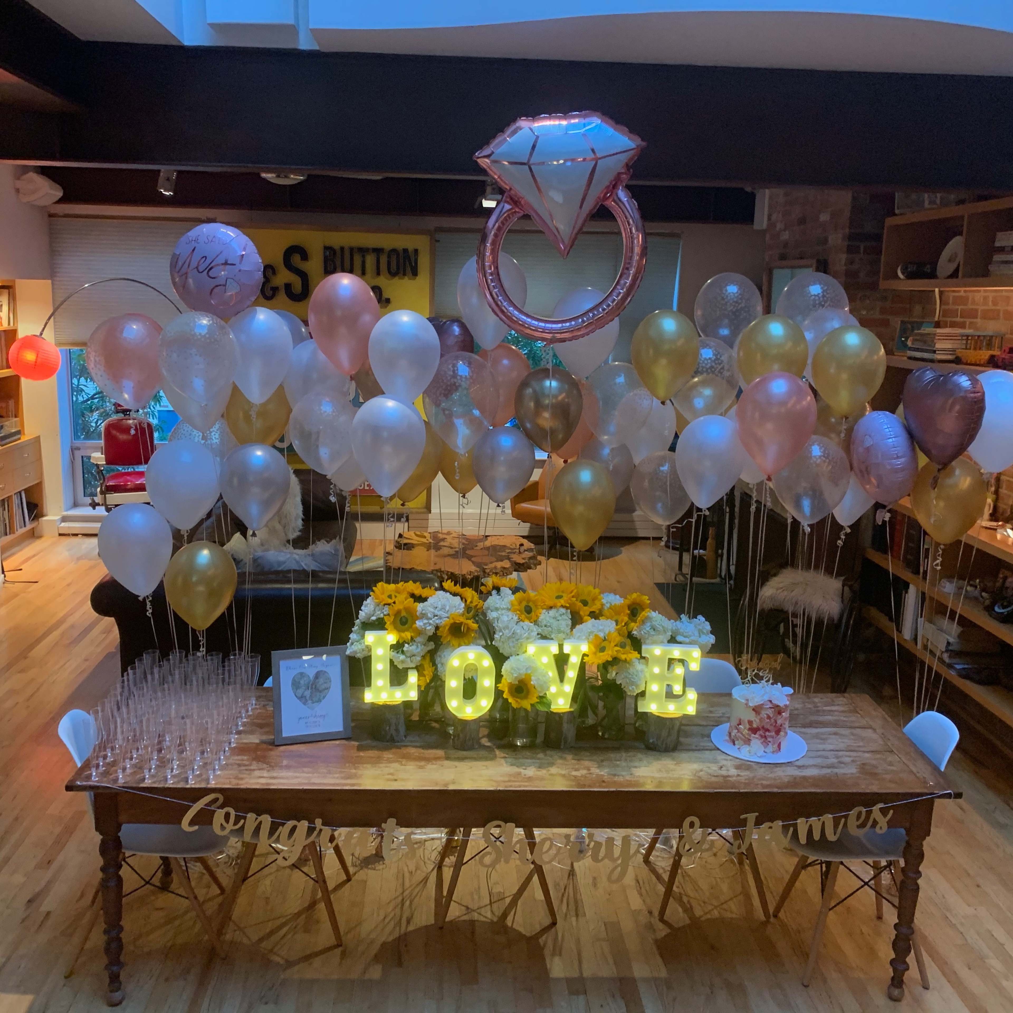 A decorated table features a large "LOVE" sign, numerous balloons, sunflowers, and a cake, set in a room with wooden floors and shelves filled with various items.