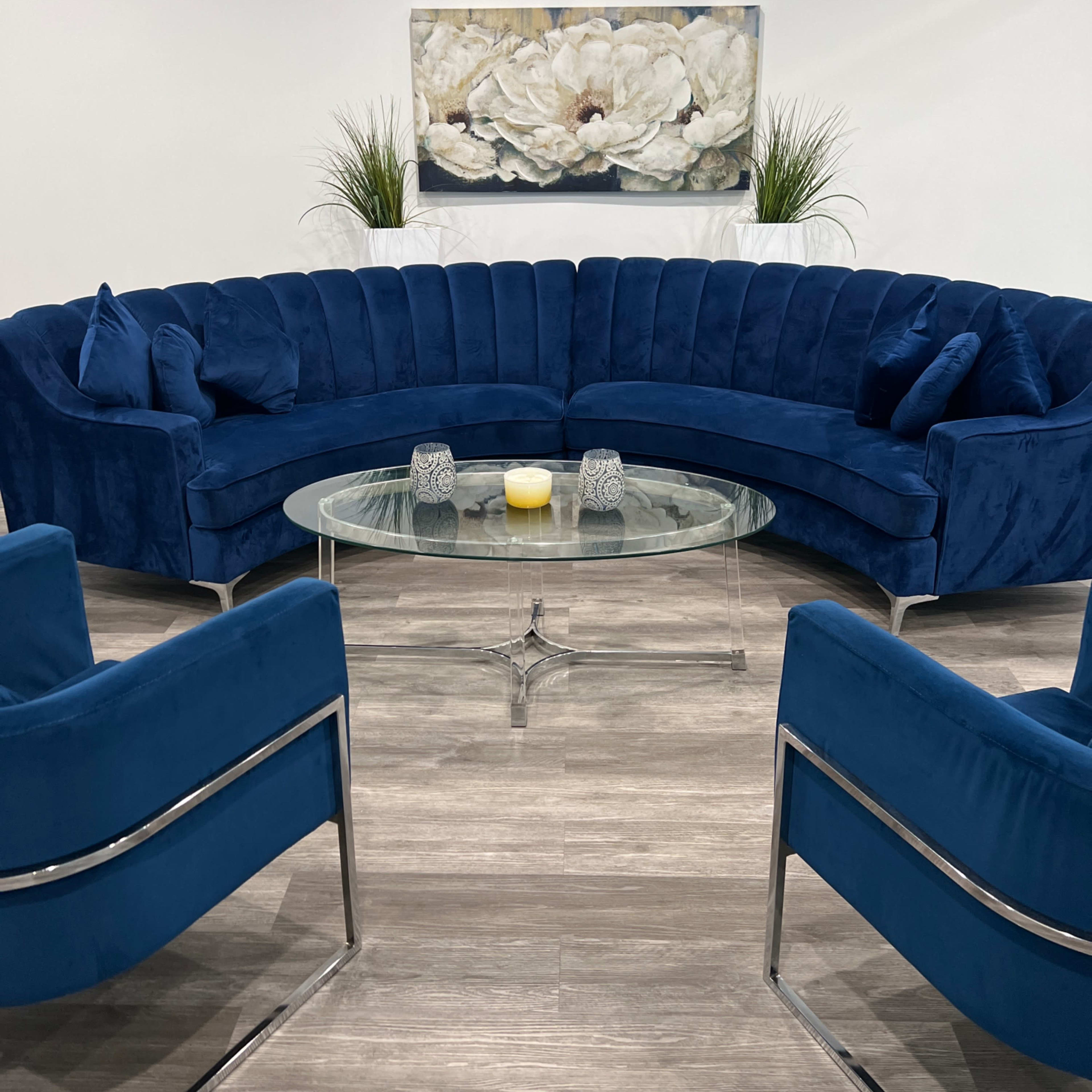 A modern lounge area with a curved blue velvet sofa, two matching armchairs, a glass coffee table, and a decorative painting of white flowers on the wall.
