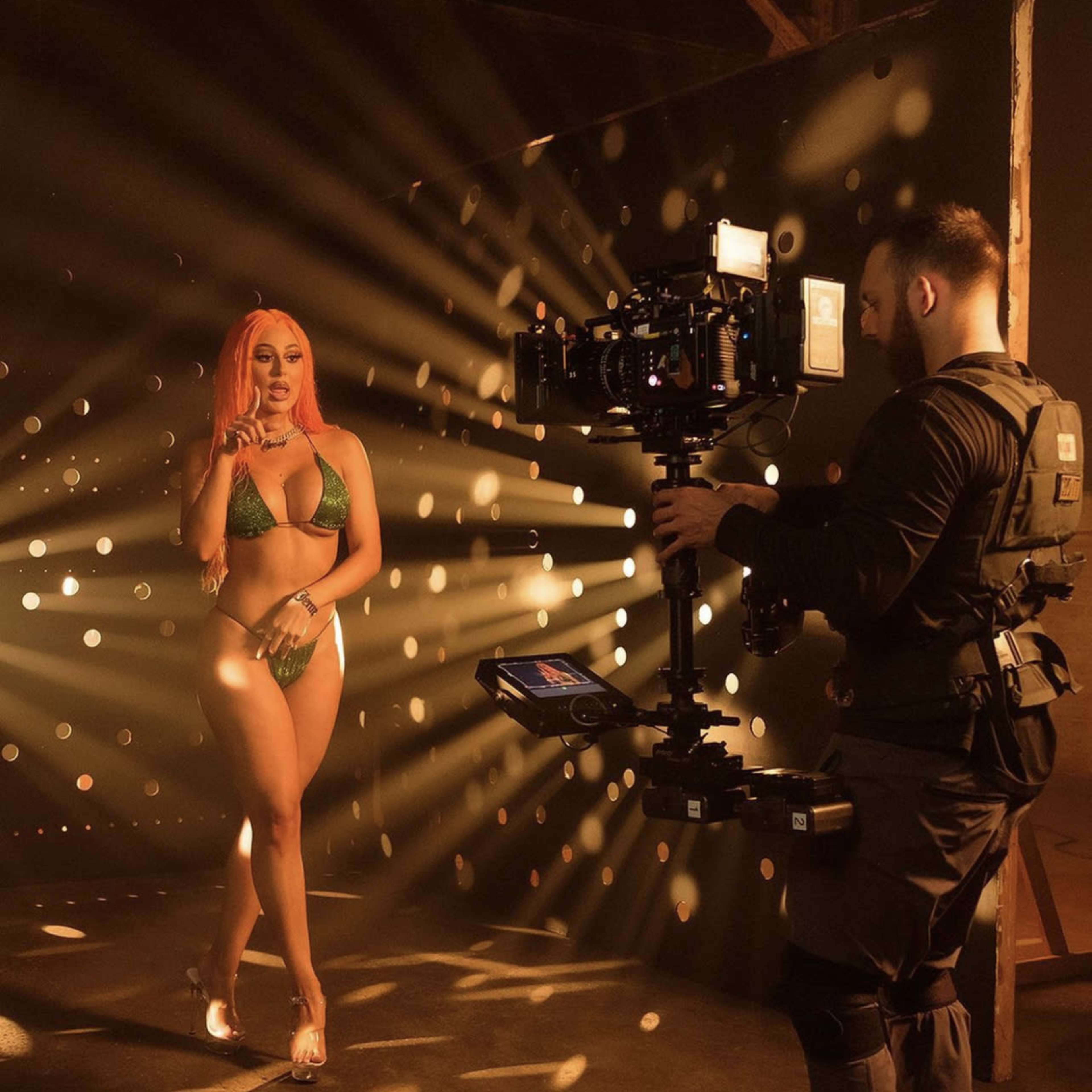 A woman in a green bikini poses in front of a camera operated by a person in a black vest, surrounded by beams of light in a dimly lit space.