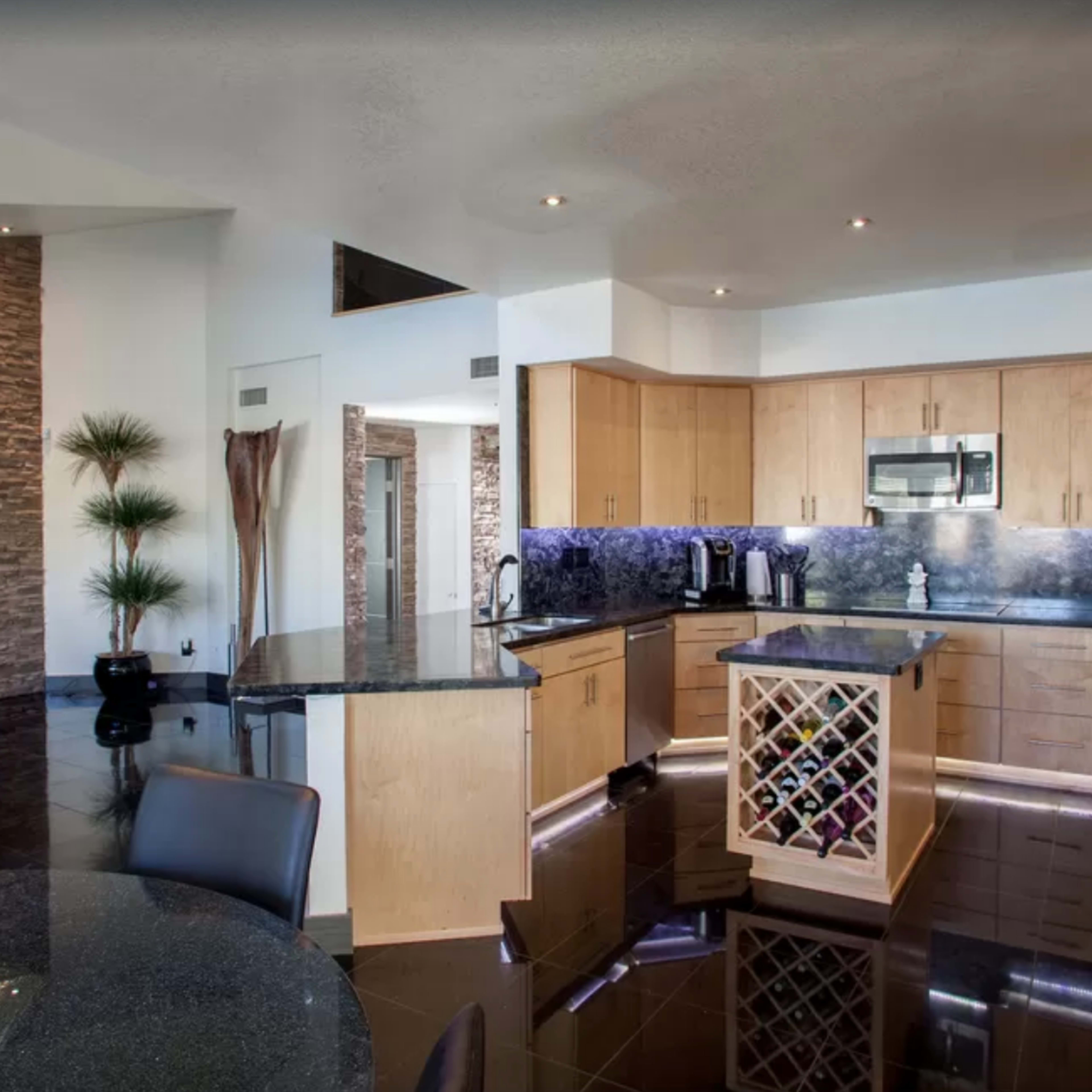 A modern kitchen with light wooden cabinetry, a dark granite countertop, and a wine rack, adjacent to a dining area with a glass table.