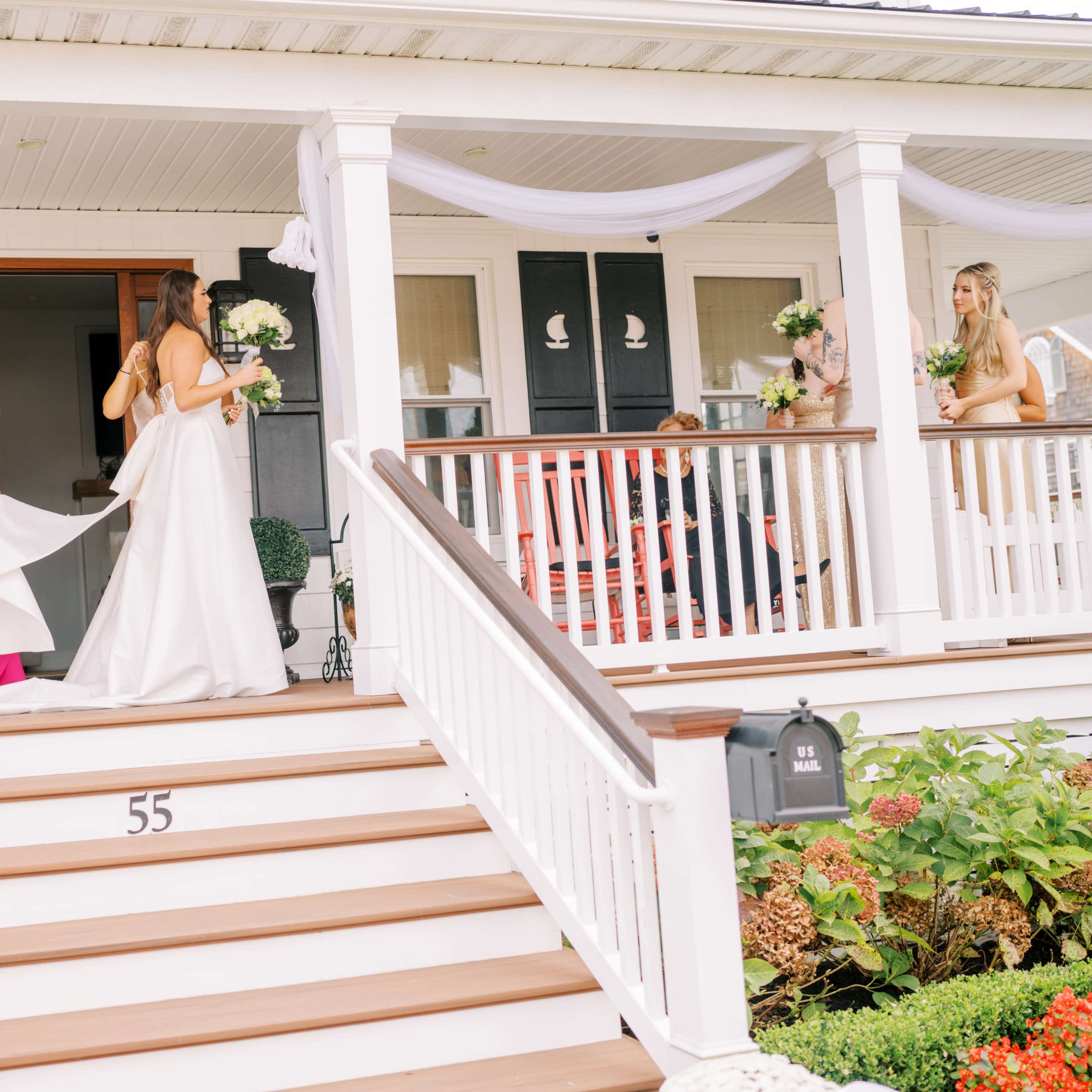 A bride in a white dress stands on the steps of a house while a woman in a pink dress helps adjust the train of her gown, and several others hold bouquets nearby.