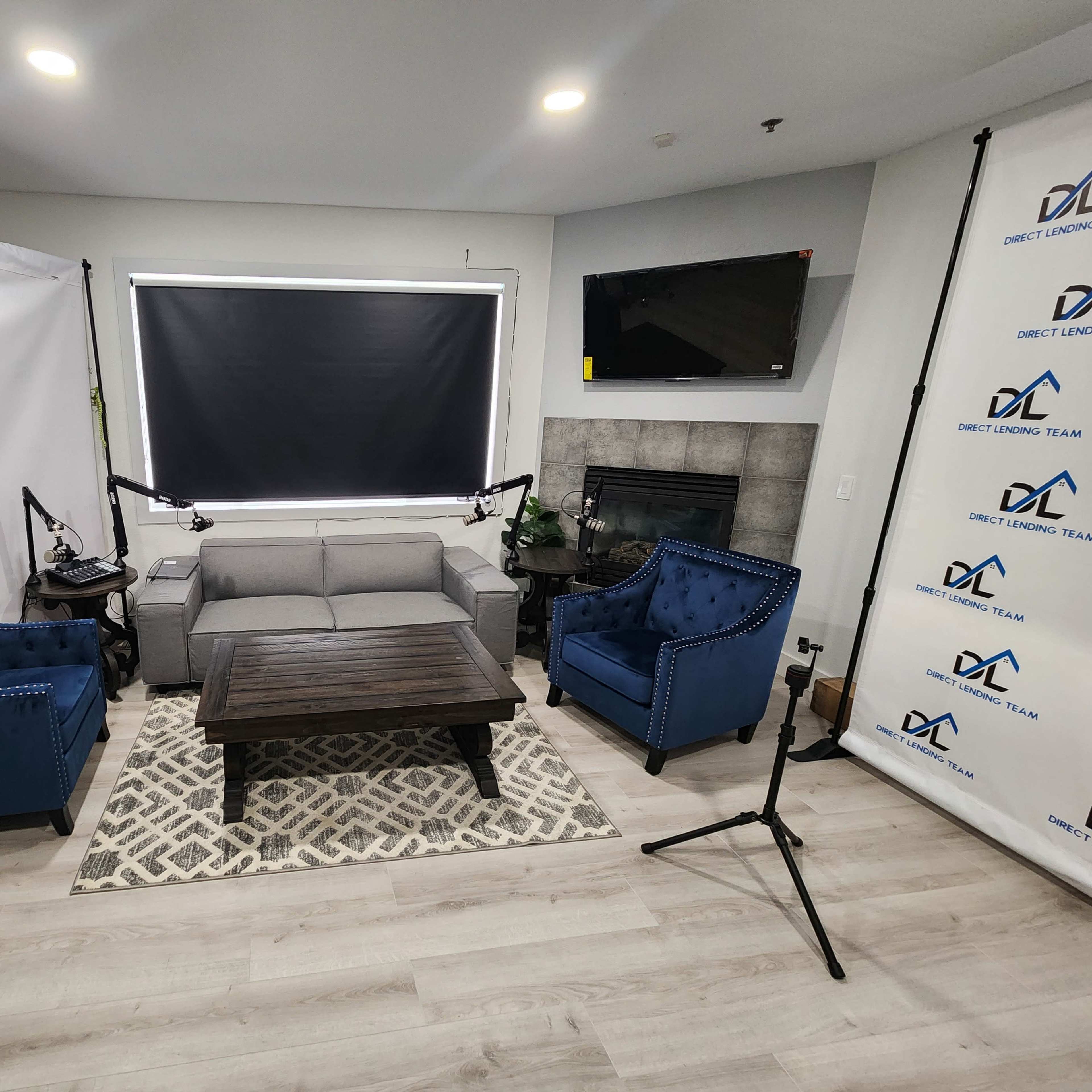 A modern living room setup with a gray sofa, two blue armchairs, a wooden coffee table, and a fireplace, all framed by a backdrop with a logo and studio lighting equipment.
