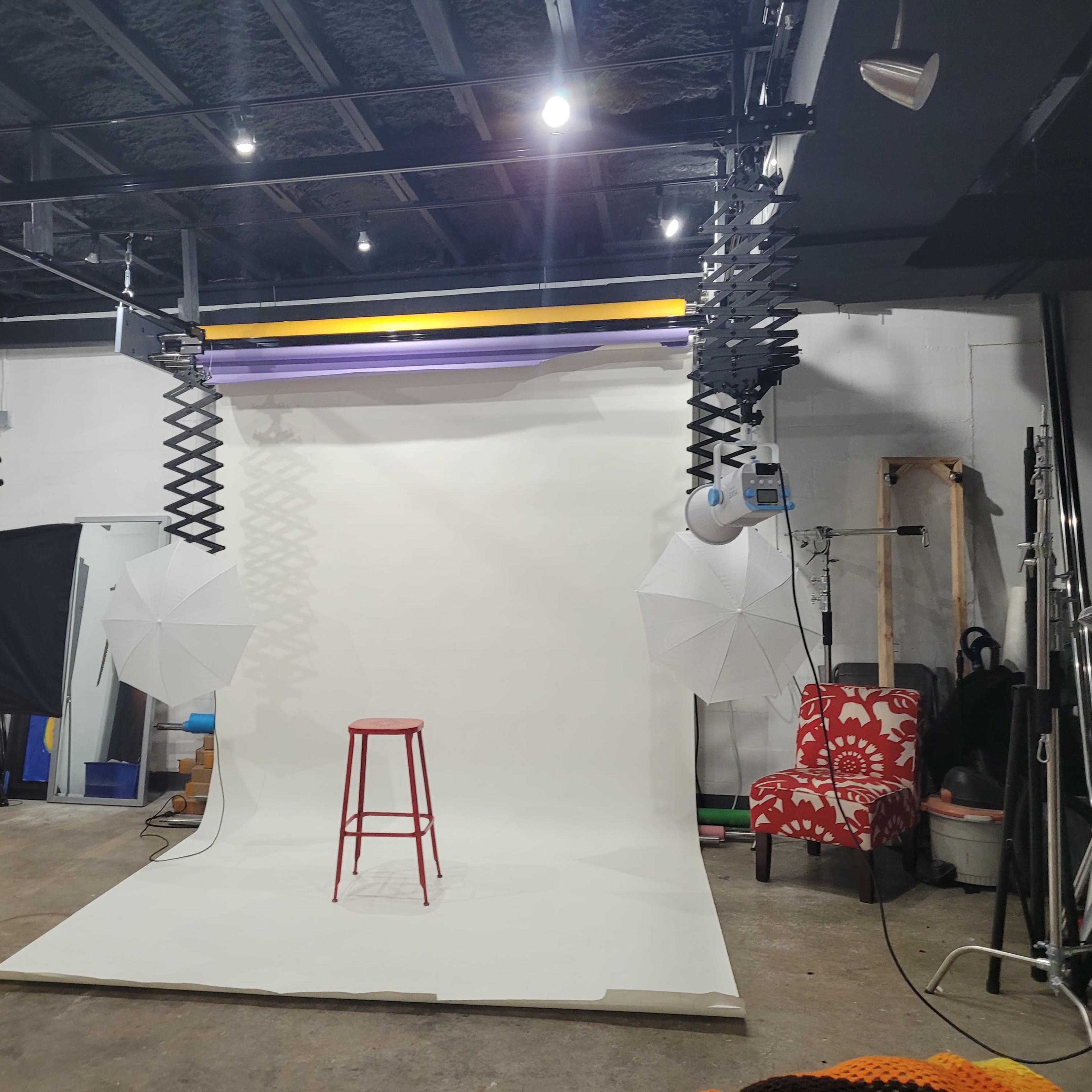 A photography studio equipped with a blank backdrop, a red stool, and lighting umbrellas set up for photo shoots.