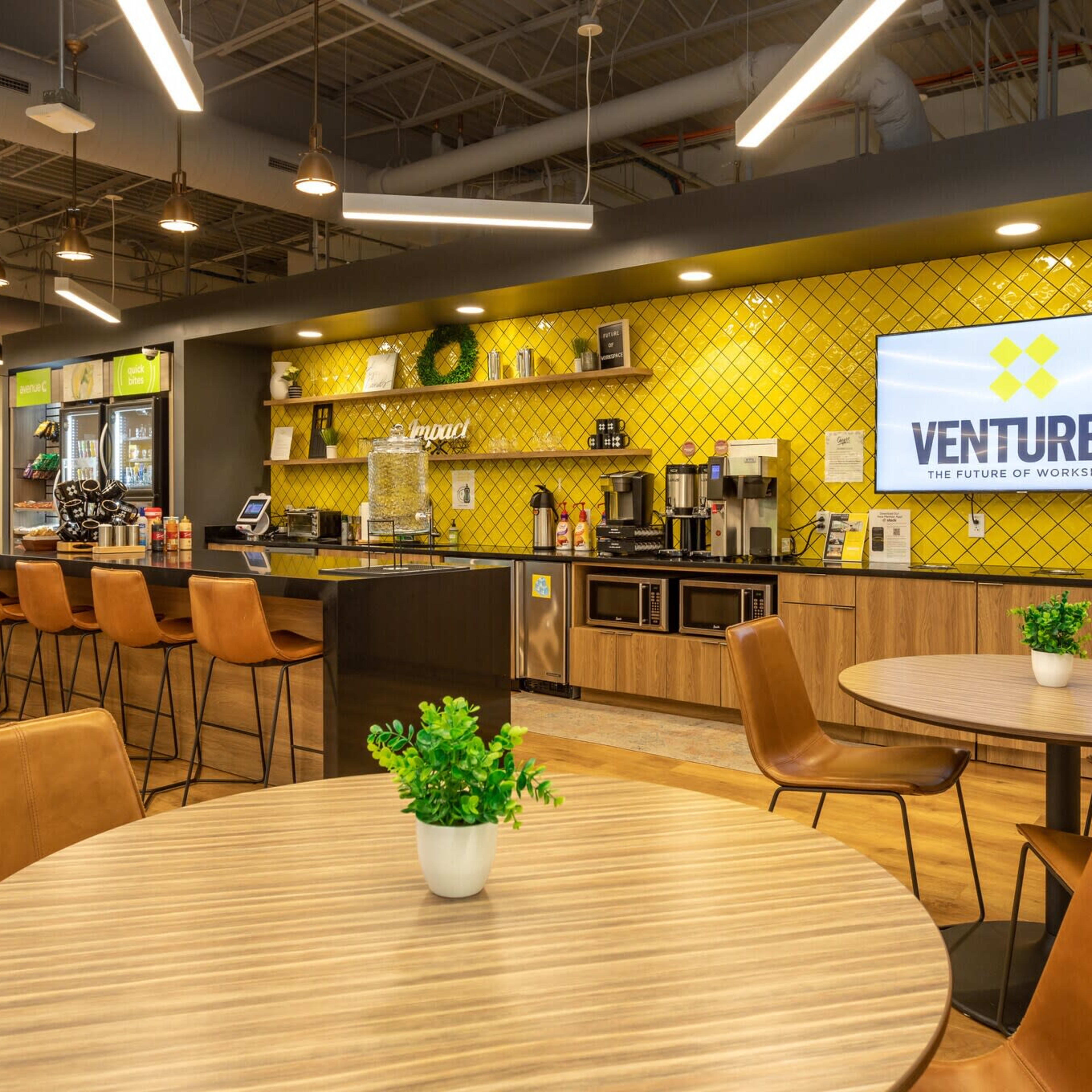A modern workspace with a kitchenette and seating area, featuring a large screen displaying "VENTURE X" and yellow tile accents.