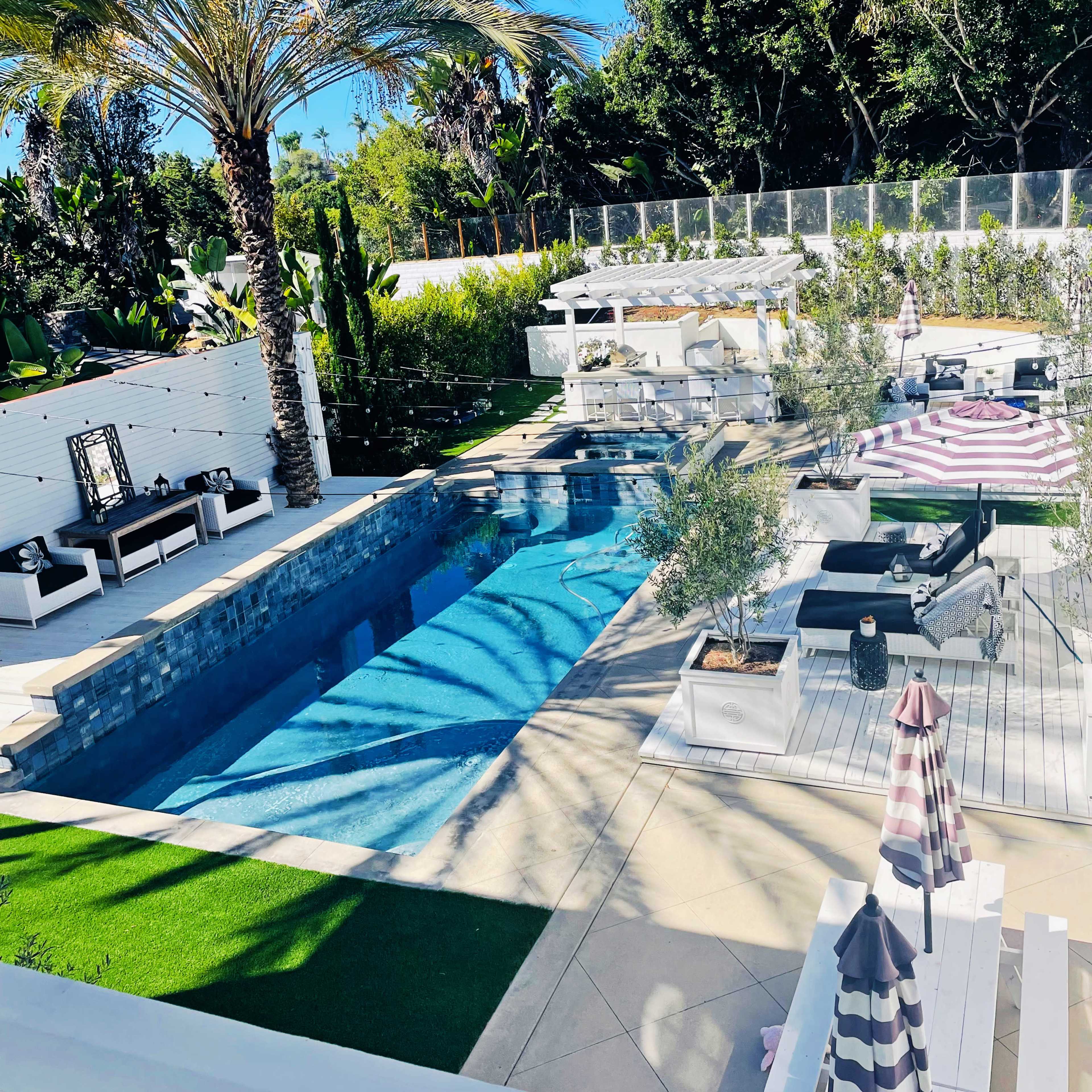 A spacious outdoor area featuring a rectangular swimming pool surrounded by lounge furniture, sunbeds, and tropical landscaping.