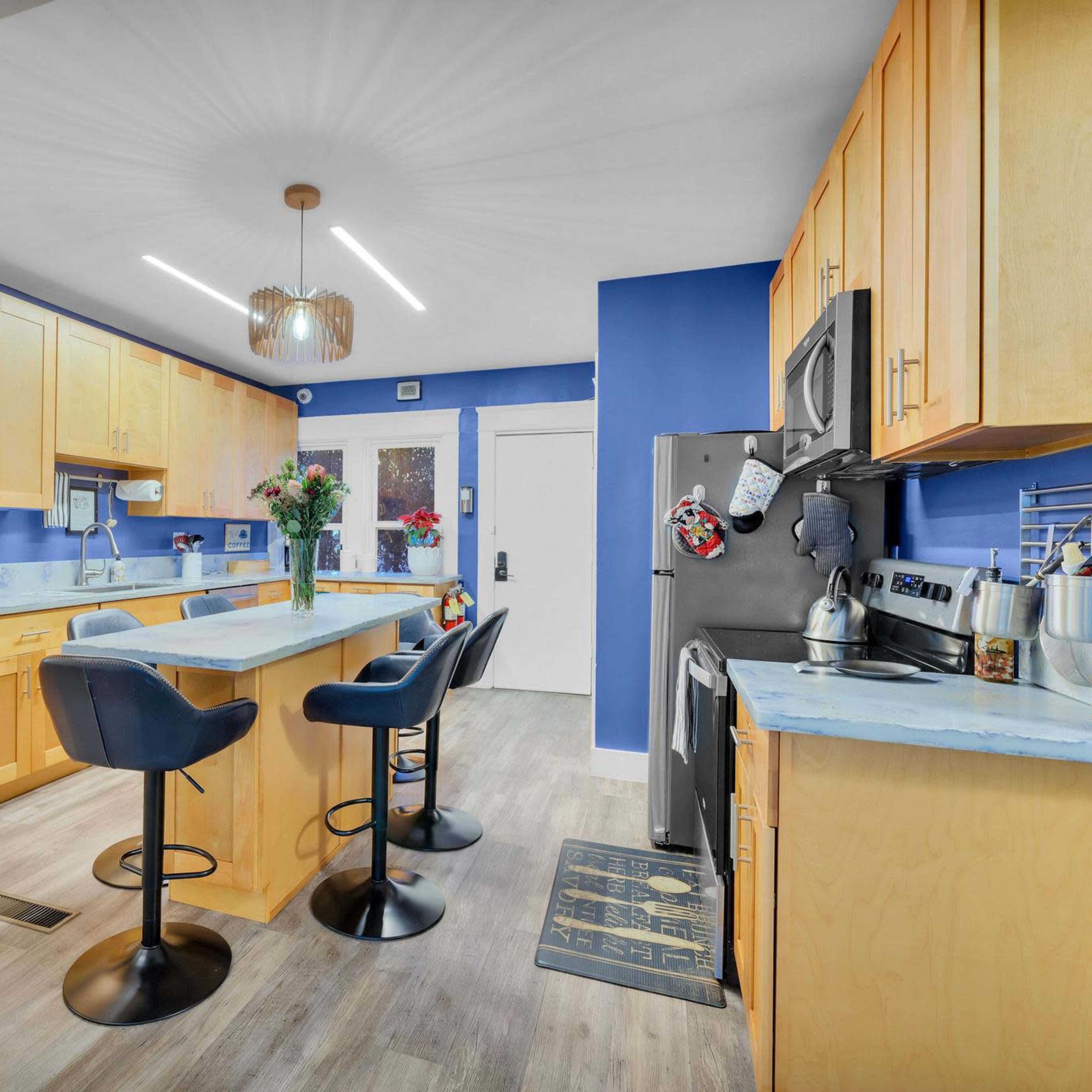 A modern kitchen with light wooden cabinets, a blue accent wall, and a central island with three black bar stools.
