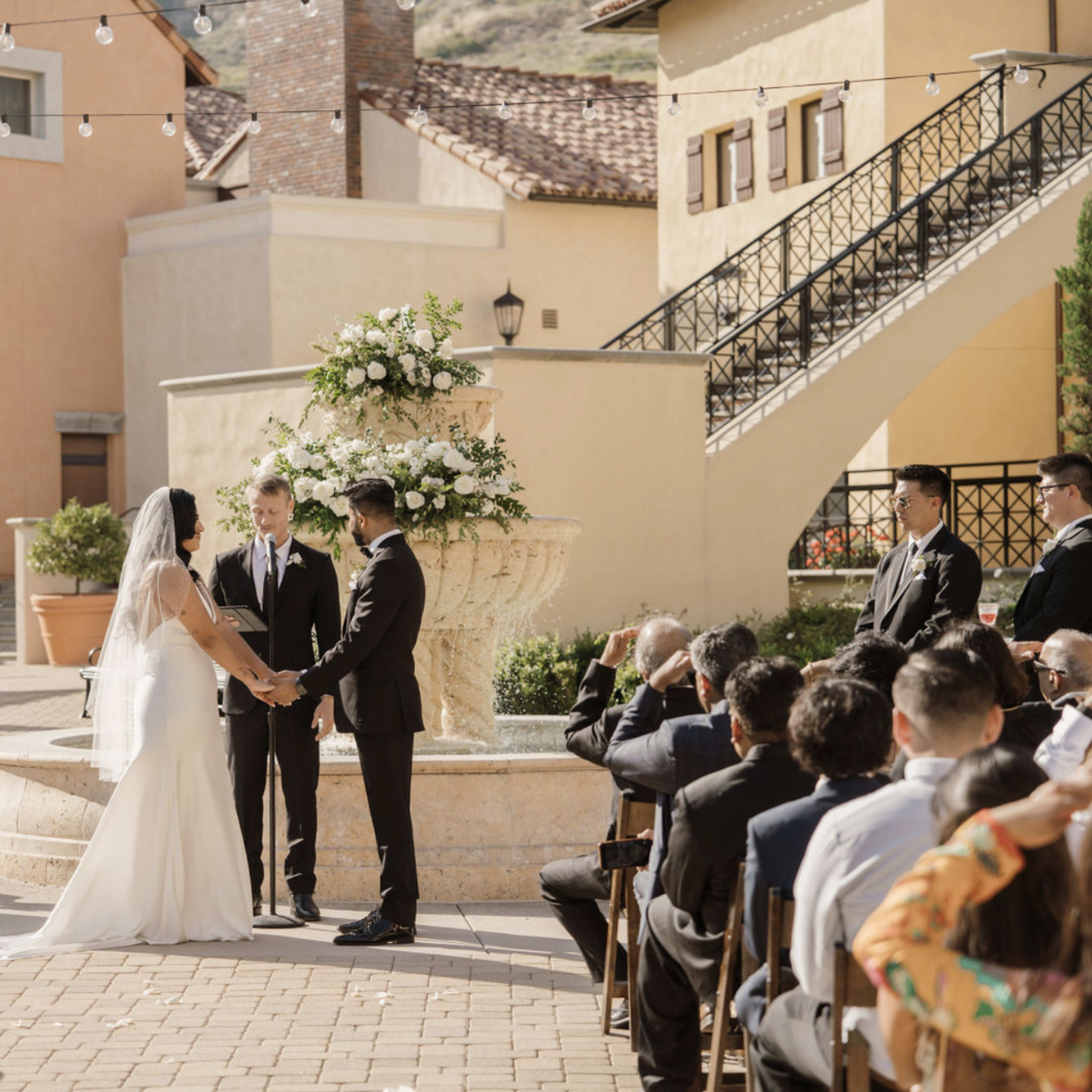 A couple exchanges vows at an outdoor wedding ceremony, surrounded by guests and decorated with floral arrangements.