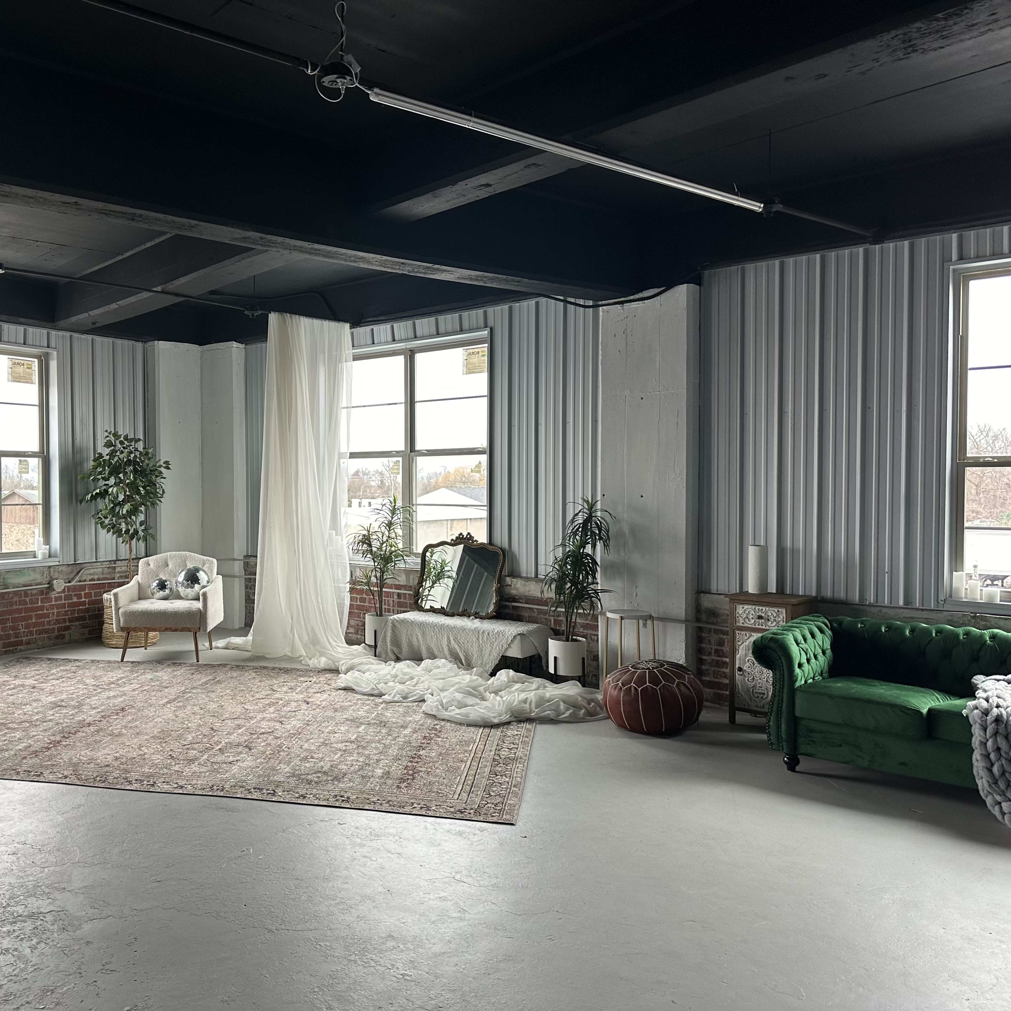 A spacious room with large windows, featuring a white sofa, a green velvet couch, a decorative rug, and various plants.