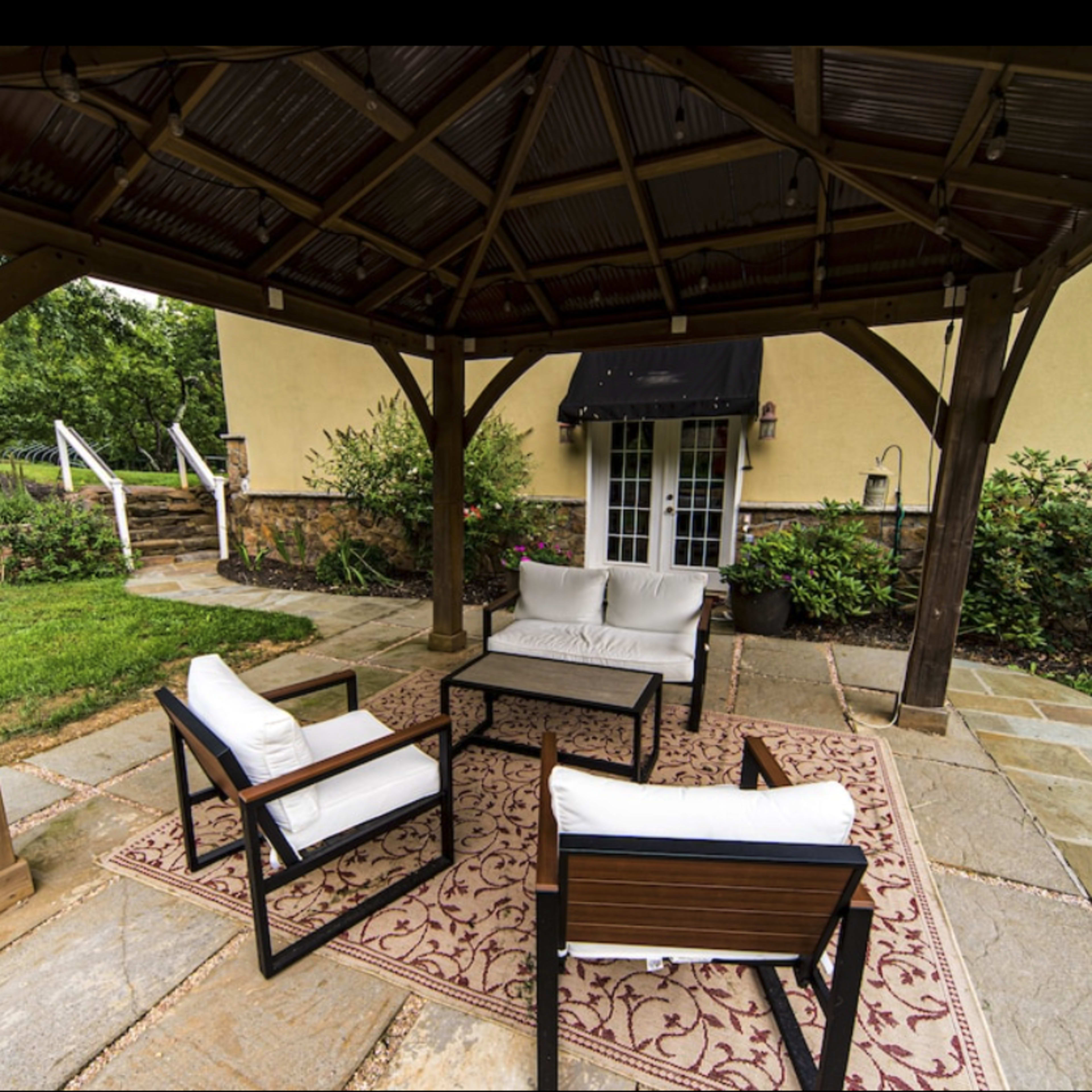 A covered outdoor seating area with four chairs and a small table on a stone patio surrounded by greenery.