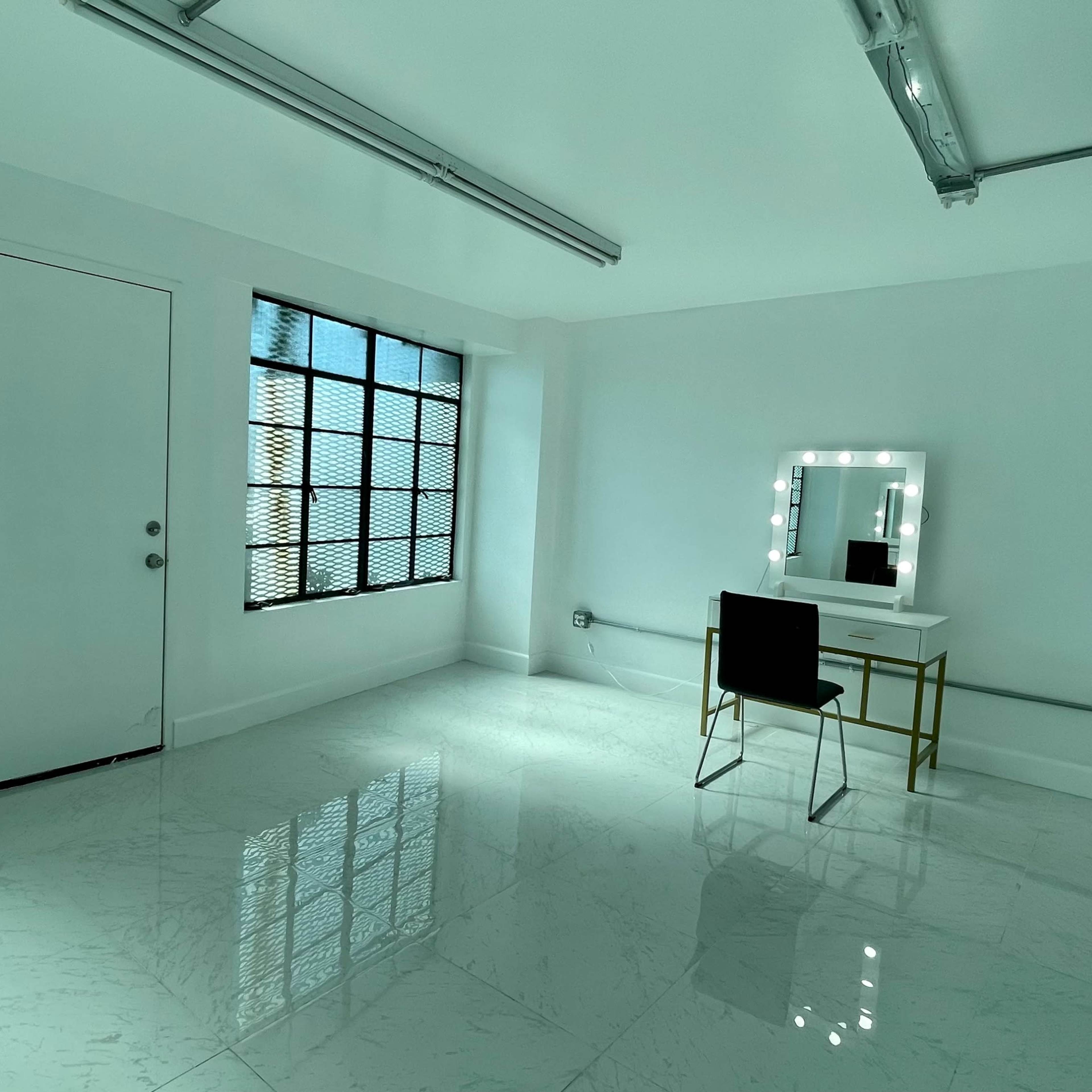 A brightly lit room with white walls, a large mirror on a vanity with lights, and a black chair, featuring two windows with metal grilles.