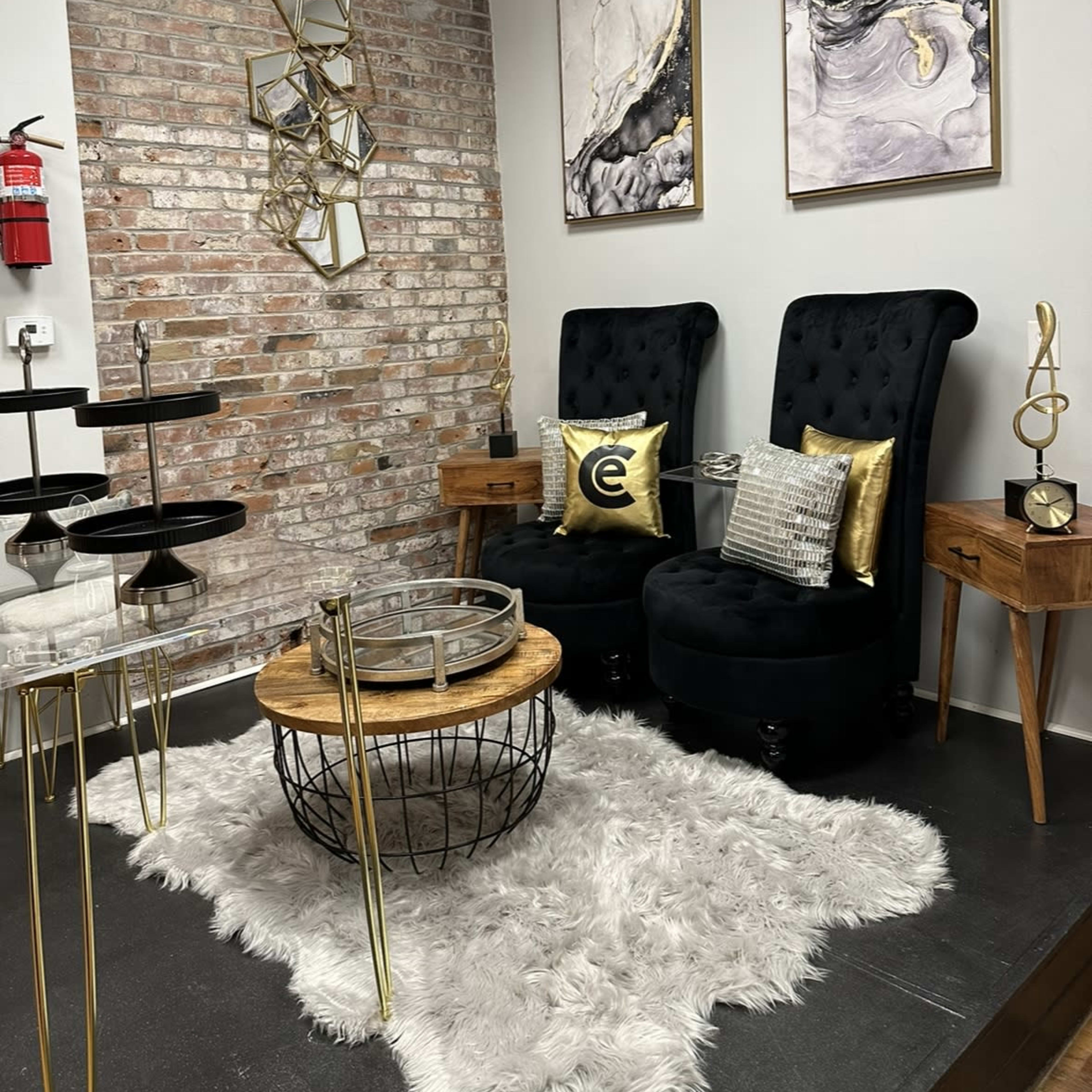 A stylish sitting area with two tufted black chairs, a round wooden coffee table, and gold accents, set against a brick wall adorned with two abstract paintings.
