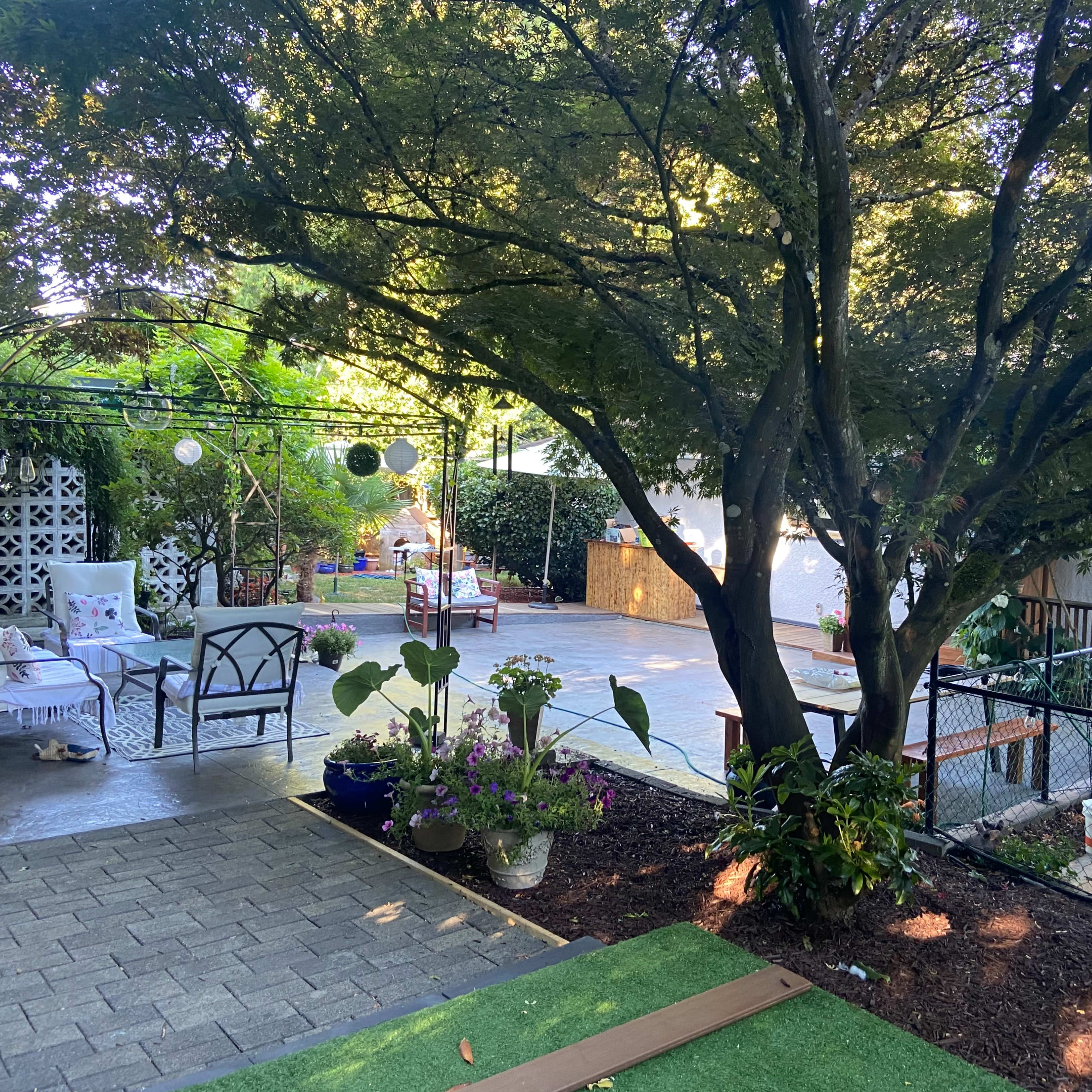A landscaped garden area with a patio, surrounded by greenery, featuring outdoor seating and a fence.