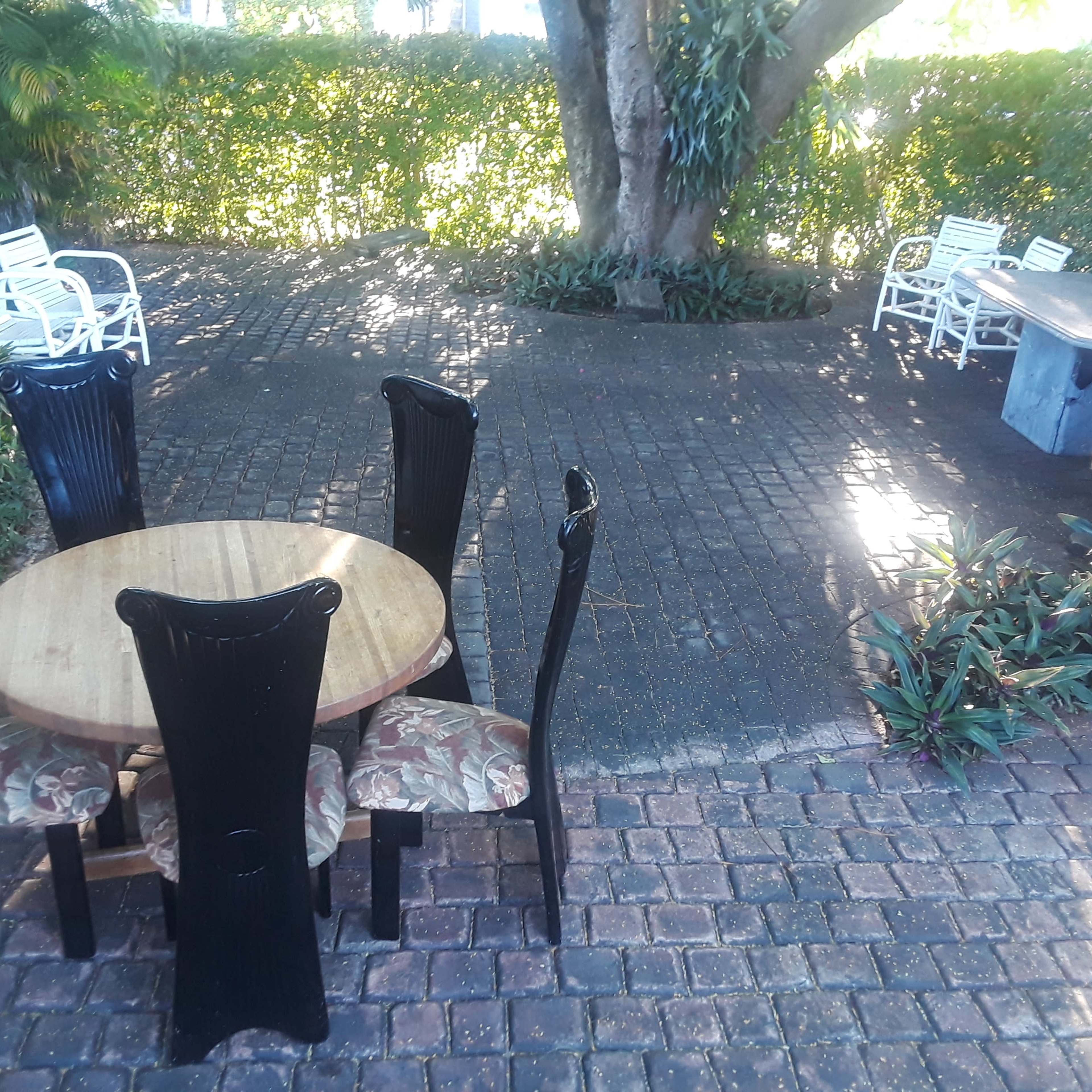 A circular wooden table with black chairs and patterned cushions is set in a stone-paved outdoor area surrounded by lush greenery.